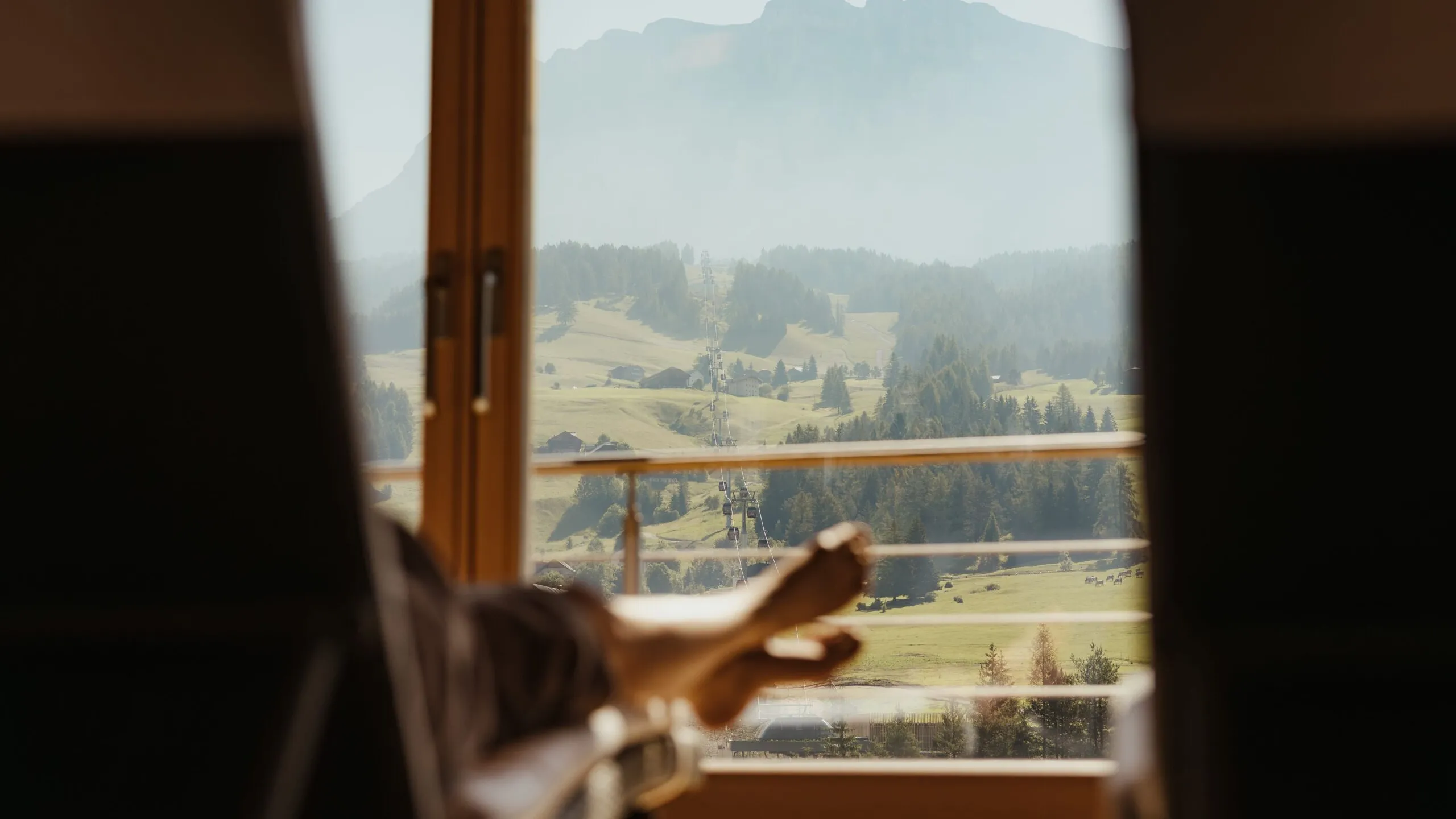 Camera al Gran Ander: persona distesa sul letto con gambe alzate, osserva dal finestrone in legno le Dolomiti, prati e boschi all’aperto.