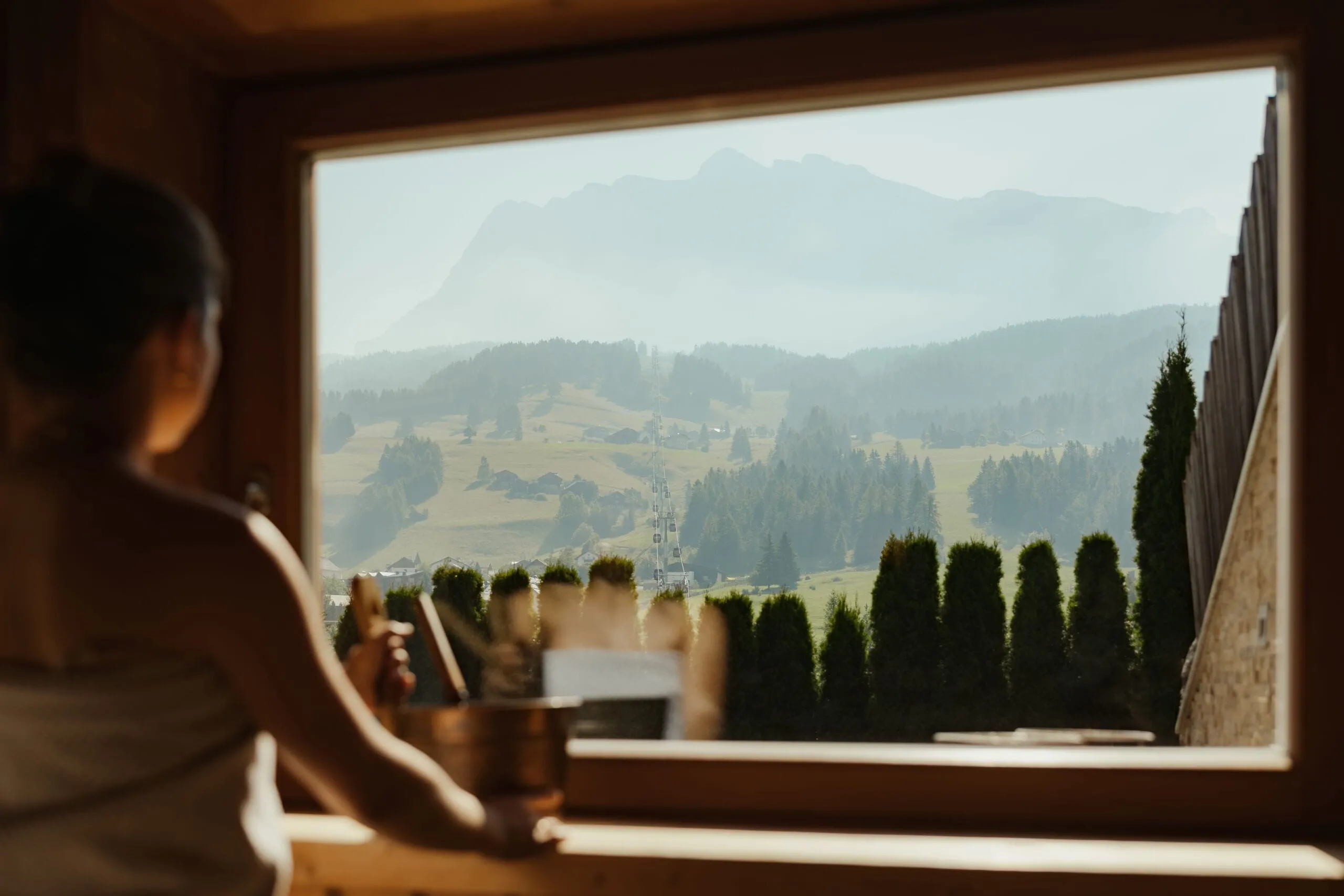 Persona seduta di schiena di fronte a una finestra in legno, osserva il paesaggio montano di Alta Badia: prati, boschi di pini e Dolomiti all’orizzonte.