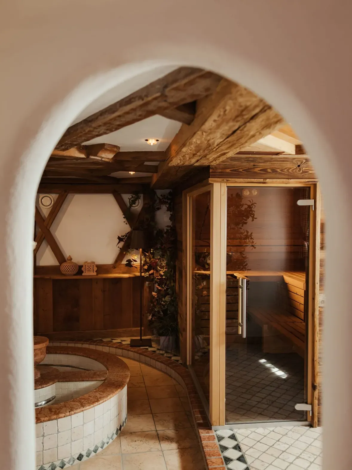 Interno spa dell'Hotel Gran Ander, Badia: arco in primo piano, vasca rotonda in mosaico, pavimenti a piastrelle, travi in legno e sauna in vetro a destra, luci calde.