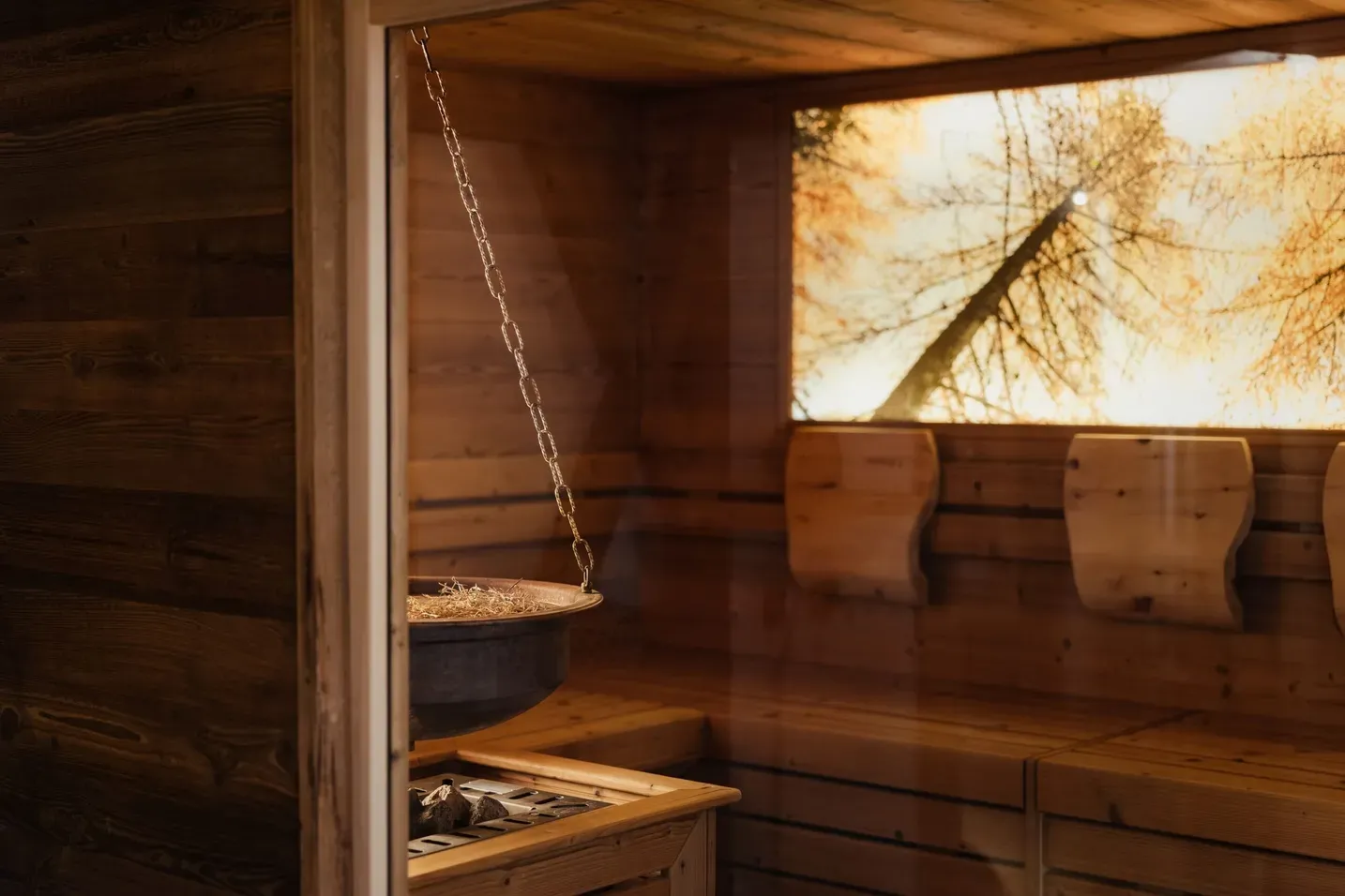 Interno di sauna in legno con braciere sospeso su catena, panche di legno e luce calda; finestra con alberi autunnali dorati.
