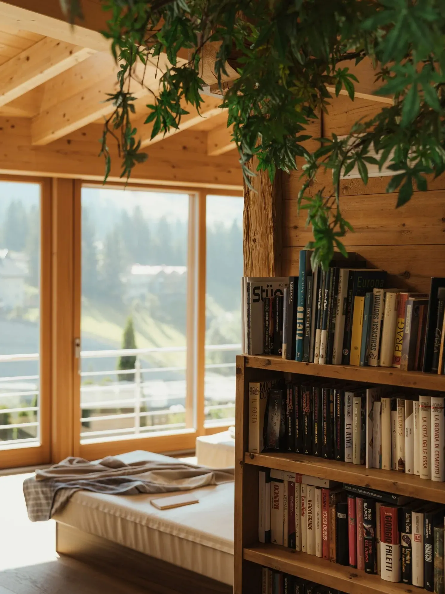 Helles Hotelzimmer im Gran Ander mit Holzwänden, Bücherregal, hängendem Grün, großen Fenstern und Blick auf die Dolomiten; Bett im Vordergrund.
