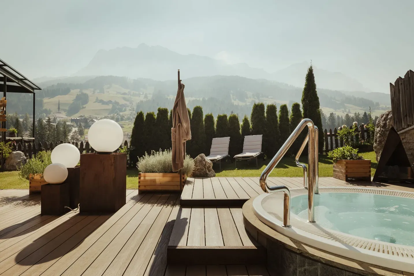 Holzdeck mit Pool und Lounge-Stühlen; runde Lichtkugeln auf Holzsäulen, grüne Hecke, Kamin rechts; Dolomiten-Bergkulisse über Alta Badia.