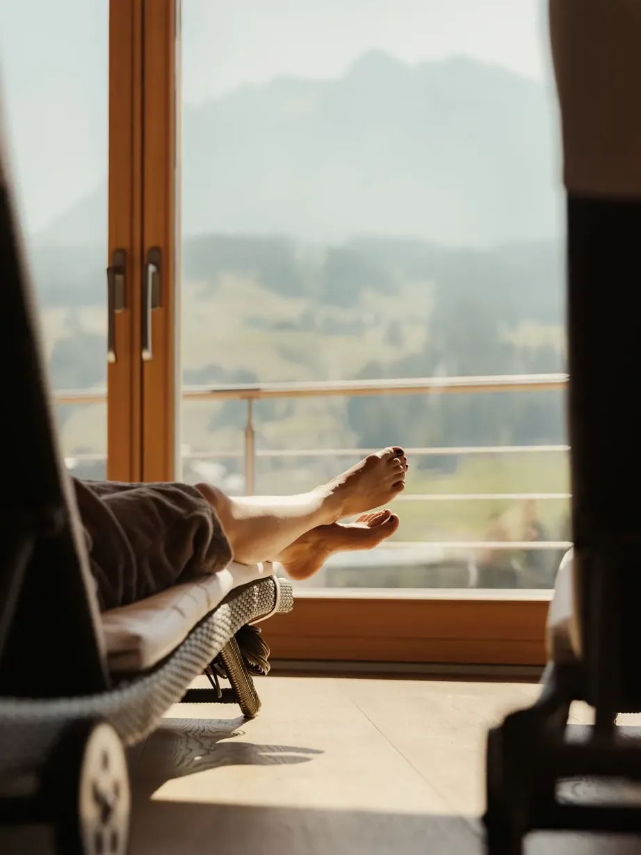 Interno di un hotel: persona distesa su una sdraio vicino a una grande finestra, piedi scoperti, coperta, luce calda e vista sfocata sulle Dolomiti.