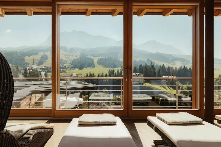 Terrazza interna con lettini di legno e grandi finestre, vista sulle Dolomiti tra prati, alberi e colline.