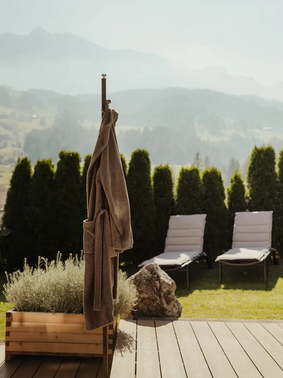 Terrazza esterna in Alta Badia, Dolomites: accappatoio marrone appeso a un palo, due lettini bianchi, aiuola in legno con lavanda e rocce, sfondo di montagne dolomitiche.