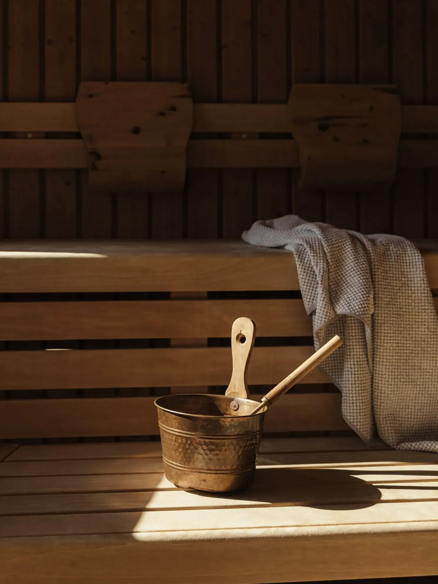 Sunlit wooden sauna interior with stacked benches, a copper bucket and ladle on the bench, and a gray towel draped nearby.