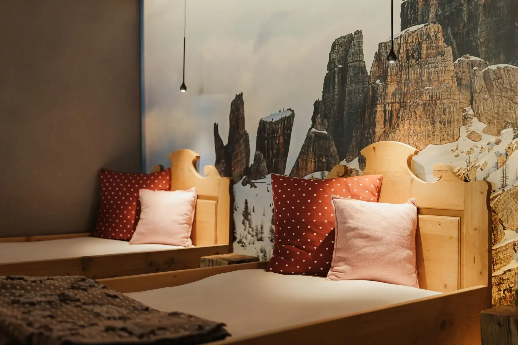 Camera da letto dell'Hotel Gran Ander, stile chalet: due letti in legno con cuscini rossi a pois e rosa, murale di rocce alpine e luci a pendente.