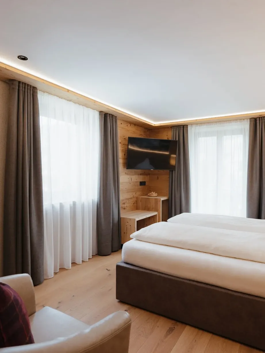 Helles Hotelzimmer mit Holzverkleidung, zwei zusammengeschobene Betten, weiße Bettwäsche, Sofa mit roten Plaid-Kissen, TV an der Wand, Fenster mit Vorhängen.