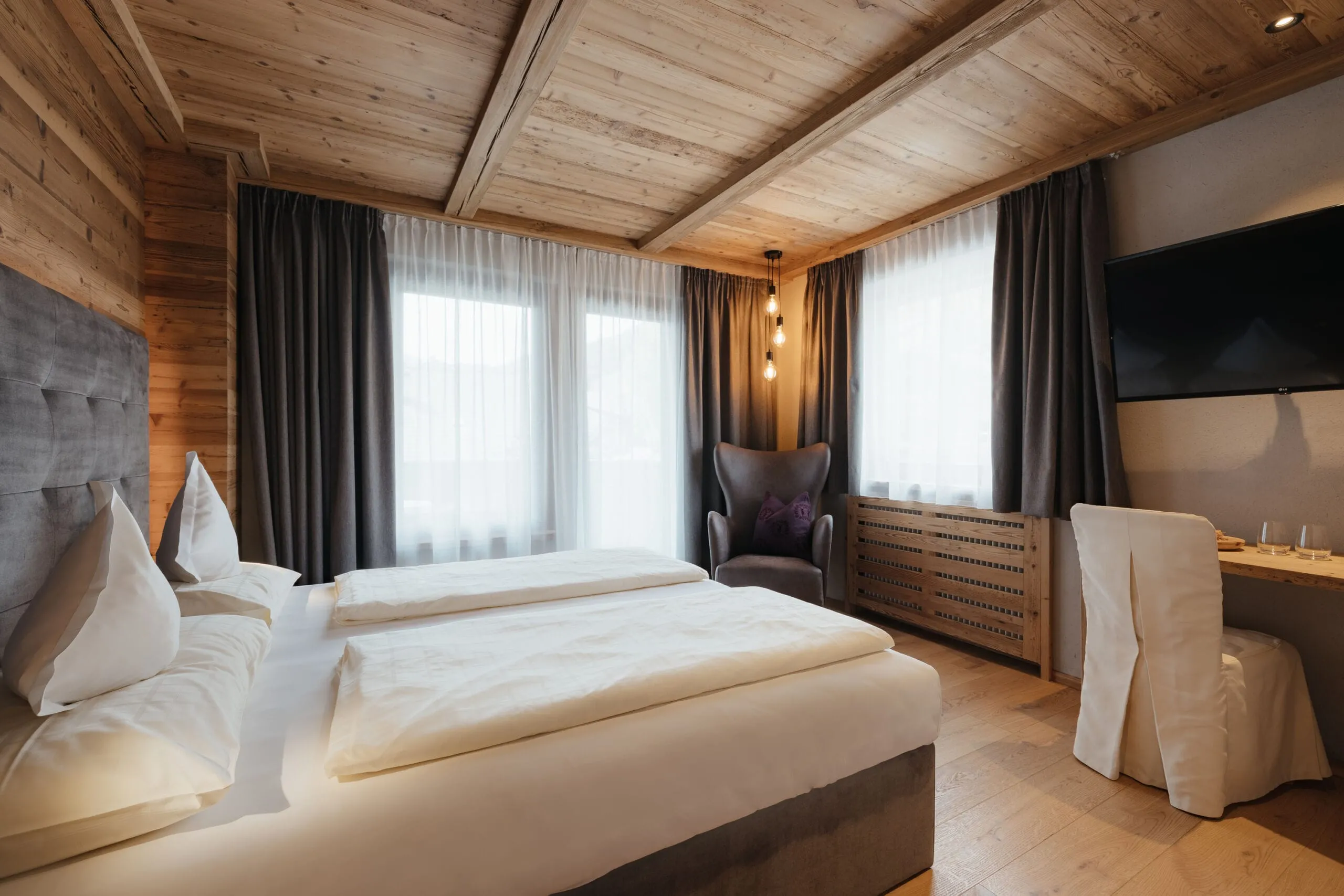 Hotelzimmer im Holzstil: Großes Bett mit weißer Wäsche, graues Kopfteil, dunkle Vorhänge, purpurfarbener Polsterstuhl, Hängeleuchten, Schreibtisch mit Stuhl und TV.