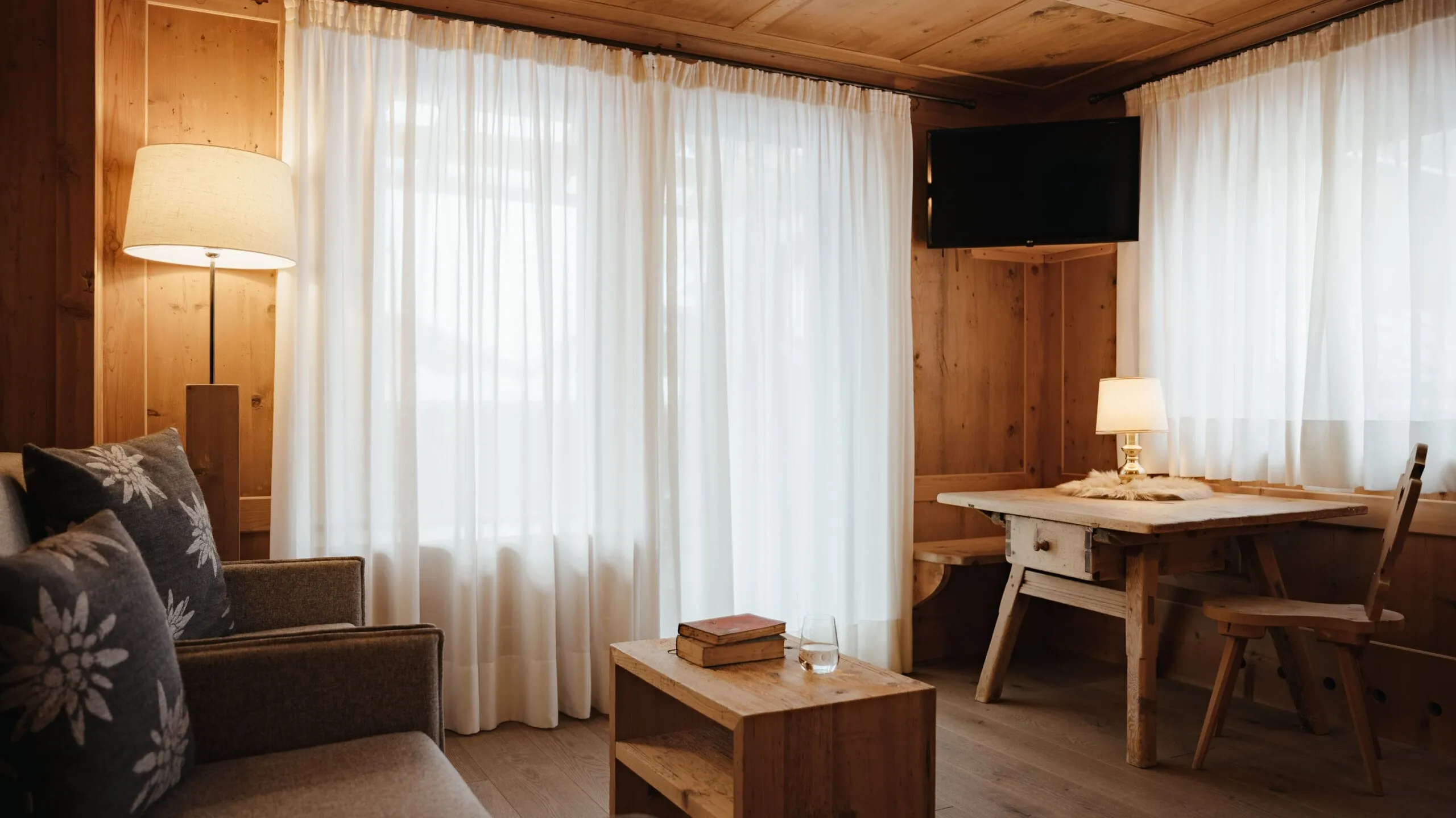Rustic wood-paneled hotel living area with sheer white curtains, a sofa with floral pillows, a wooden coffee table with books, and a dining desk with chairs.