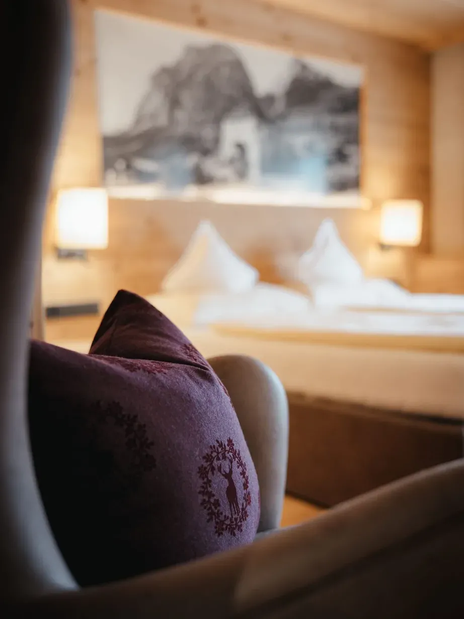 Violettes Zierkissen im Vordergrund eines Hotelzimmers im Hotel Gran Ander, Badia, Alta Badia, Dolomiten; Bett, Holzwand und warme Lampen im Hintergrund.