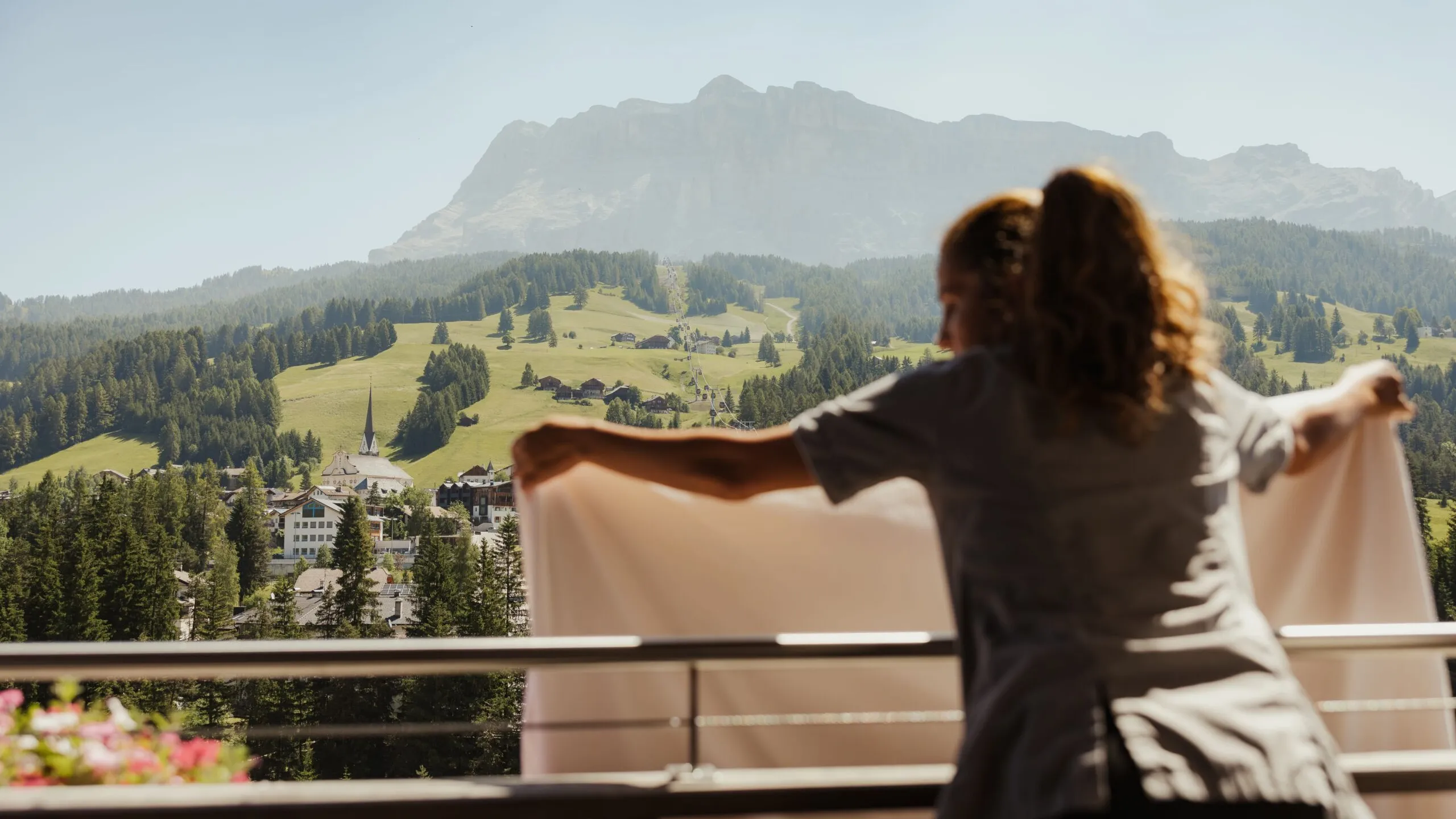 Persona vista di schiena sul balcone, braccia tese con un telo bianco; sfondo Dolomiti, pendii verdi e un villaggio con chiesa.