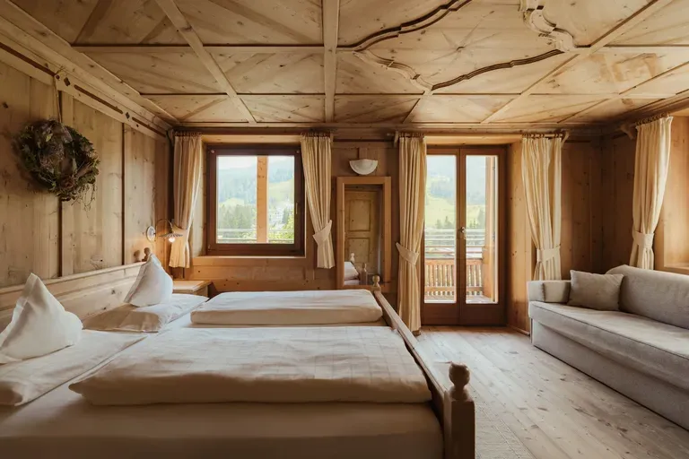 Helles Holzzimmer im alpinen Stil mit Bett, Sofa, großen Fenstern und Balkonzugang; Blick auf die Dolomiten.
