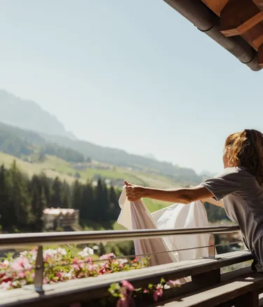 Persona su balcone di legno allunga un panno bianco; fiori sul davanzale, Alta Badia, Dolomiti sullo sfondo.