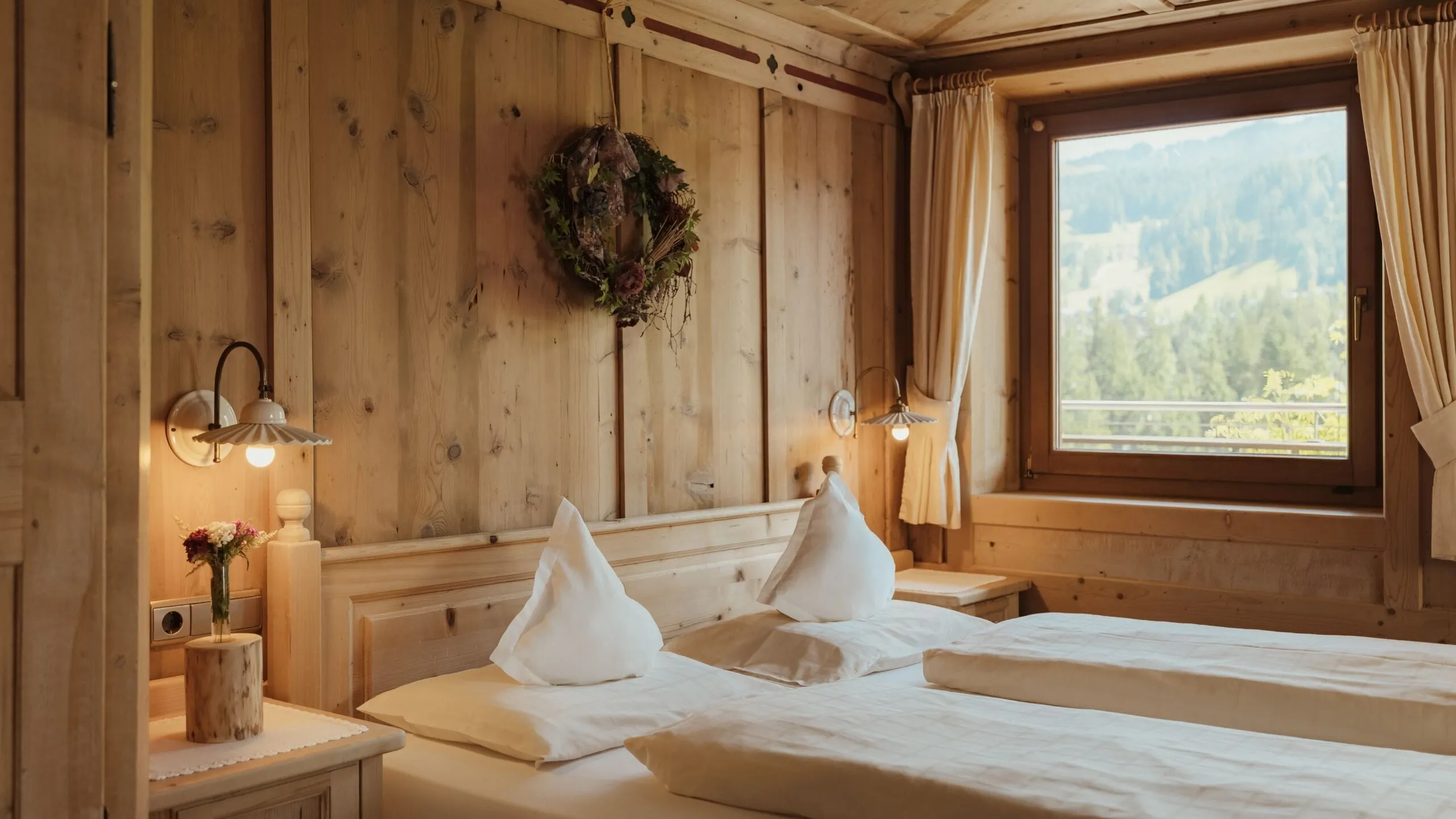 Interno in legno del Hotel Gran Ander, Badia: due letti con cuscini bianchi, luci da comodino, ghirlanda appesa, finestra con vista Dolomiti.