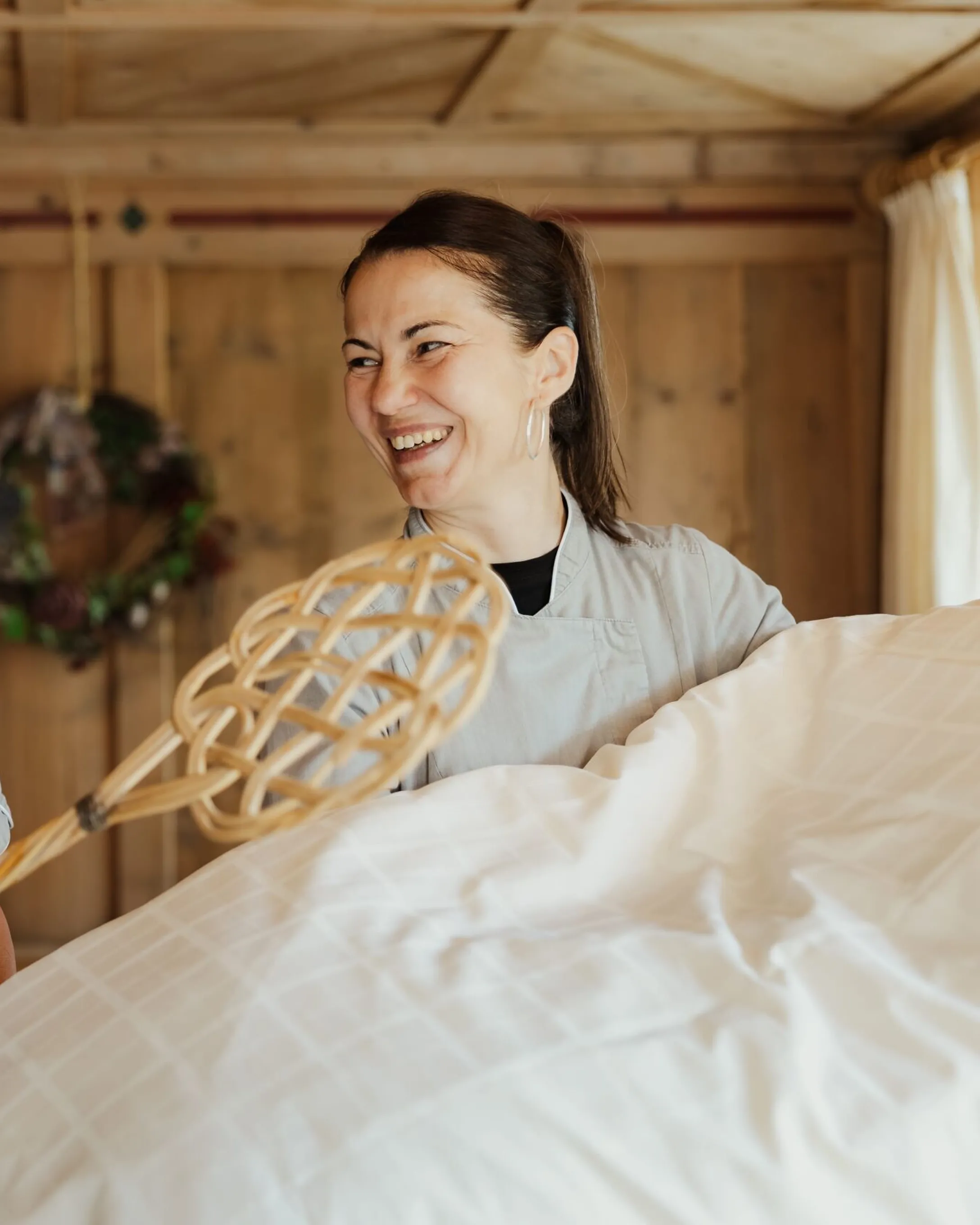 Two staff members in blue uniforms smile while making a bed with a woven bed-making tool in a warm, wood-paneled room at Hotel Gran Ander, Badia, Alta Badia.