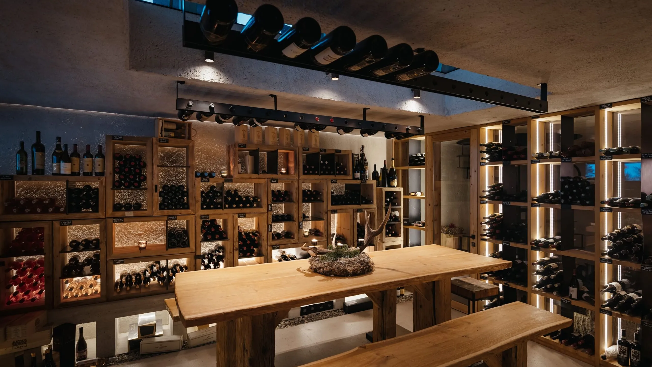 Cozy wine cellar at Hotel Gran Ander, Badia: wooden shelves stacked with wine bottles surround a central wooden table and bench, warm lighting and antler centerpiece.