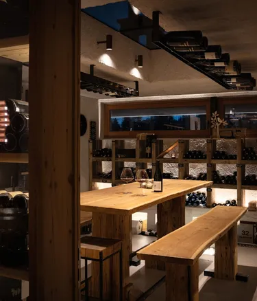 Warm wooden wine cellar and tasting room with a long table and benches, shelves of wine bottles, a bottle and glass on the table.