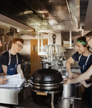 Quattro membri dello staff in cucina al Hotel Gran Ander, Badia, Alta Badia, intorno a una grande griglia nera Kamado, ridono tra utensili appesi e superfici in acciaio.