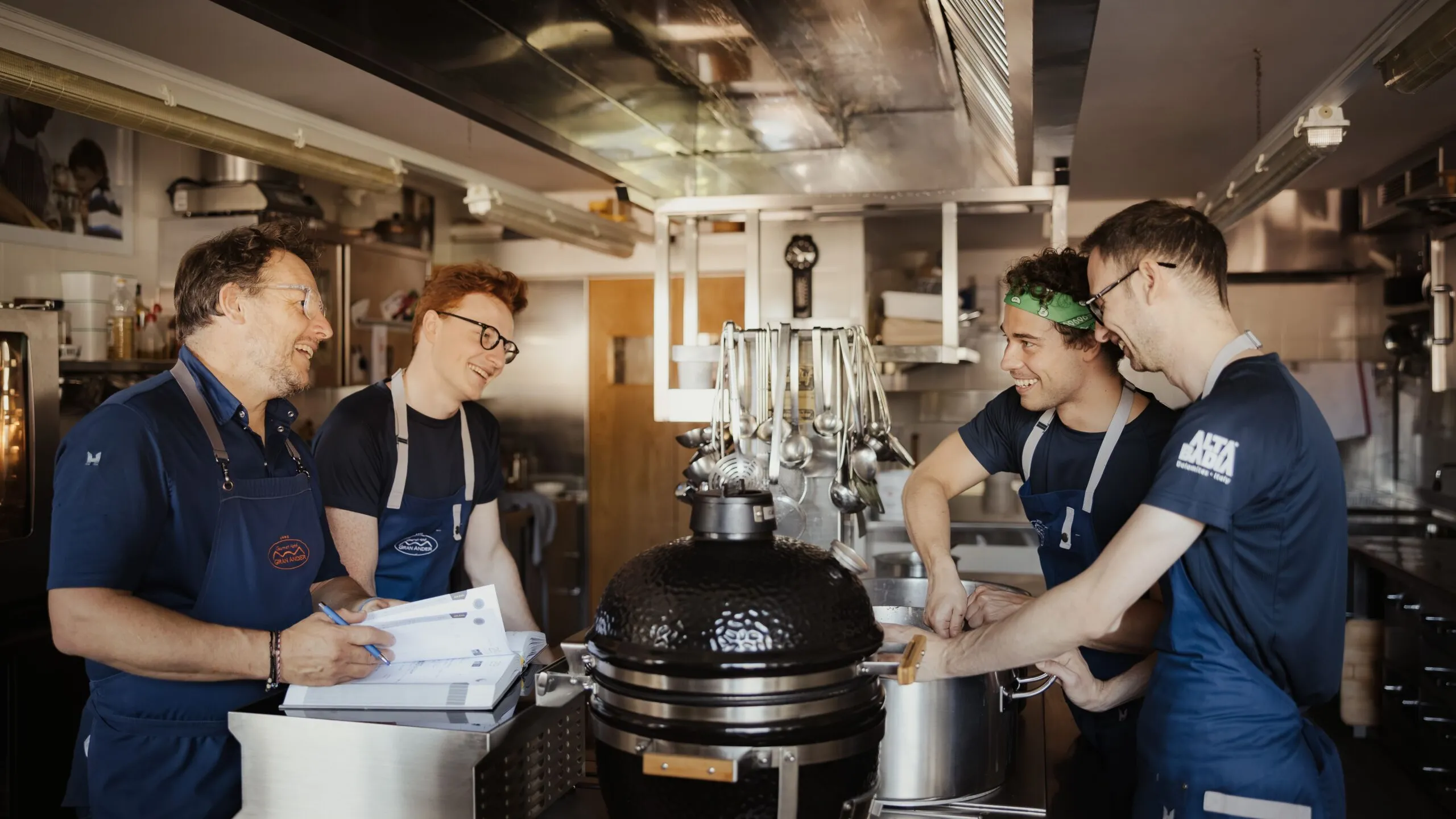 Quattro membri dello staff in cucina al Hotel Gran Ander, Badia, Alta Badia, intorno a una grande griglia nera Kamado, ridono tra utensili appesi e superfici in acciaio.