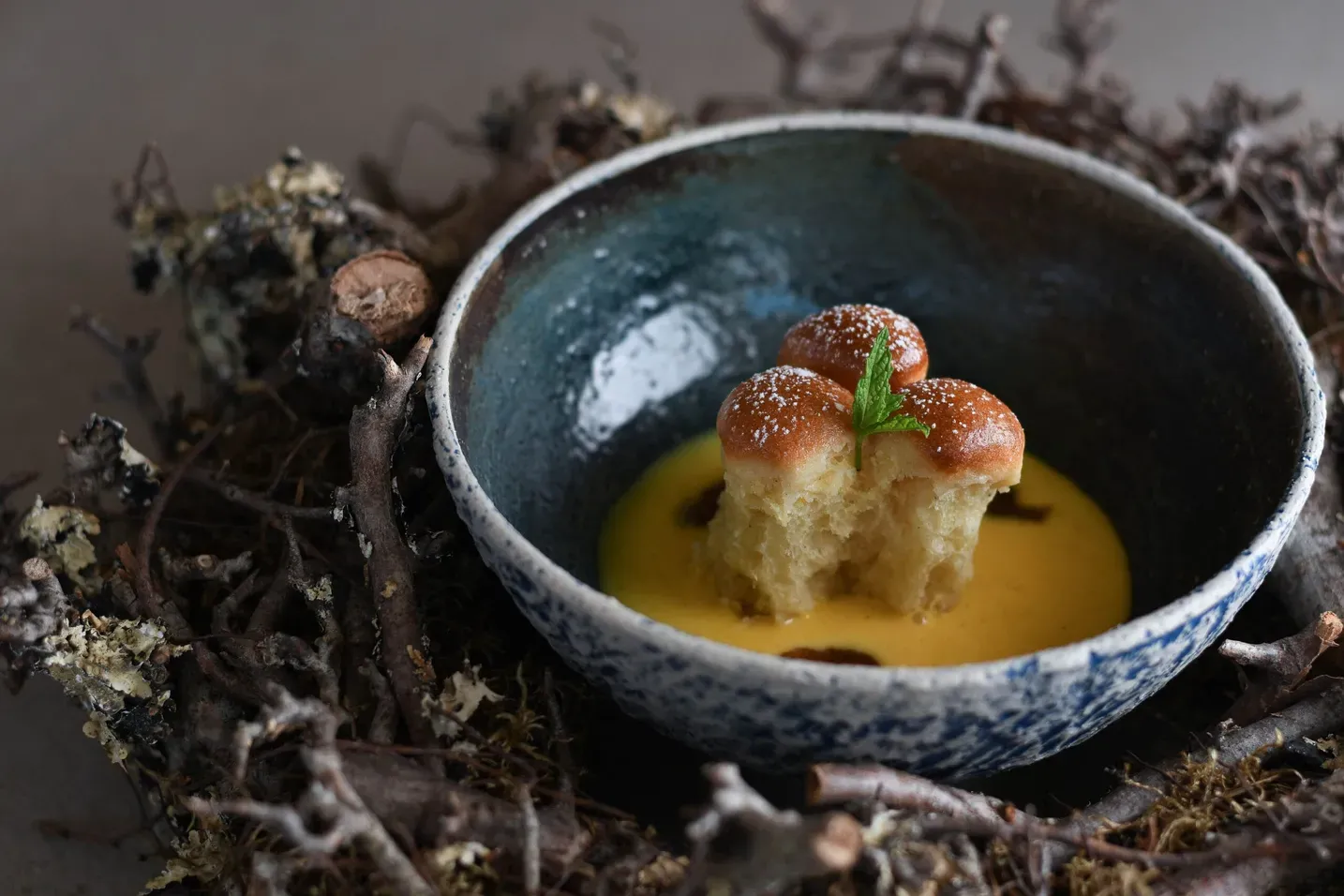 Blaues Schälchen mit gelber Sauce, drei kleine goldbraune Brioche-Stückchen mit Minze, in einem Nest aus Zweigen und Moos (Alta Badia, Dolomiten).