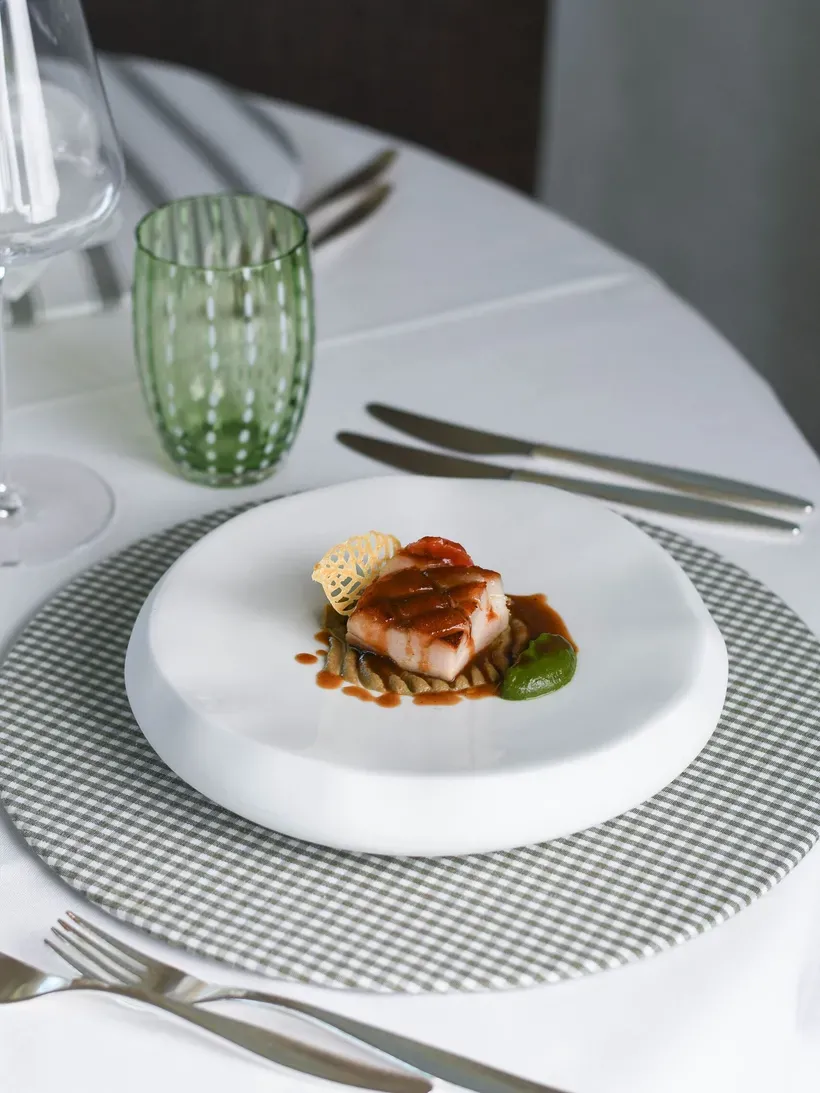 Feines Gourmetgericht: gebratener Fisch mit brauner Sauce, grünem Dip und knusprigem Crisp auf einer runden Beilage, serviert auf weißem Teller.