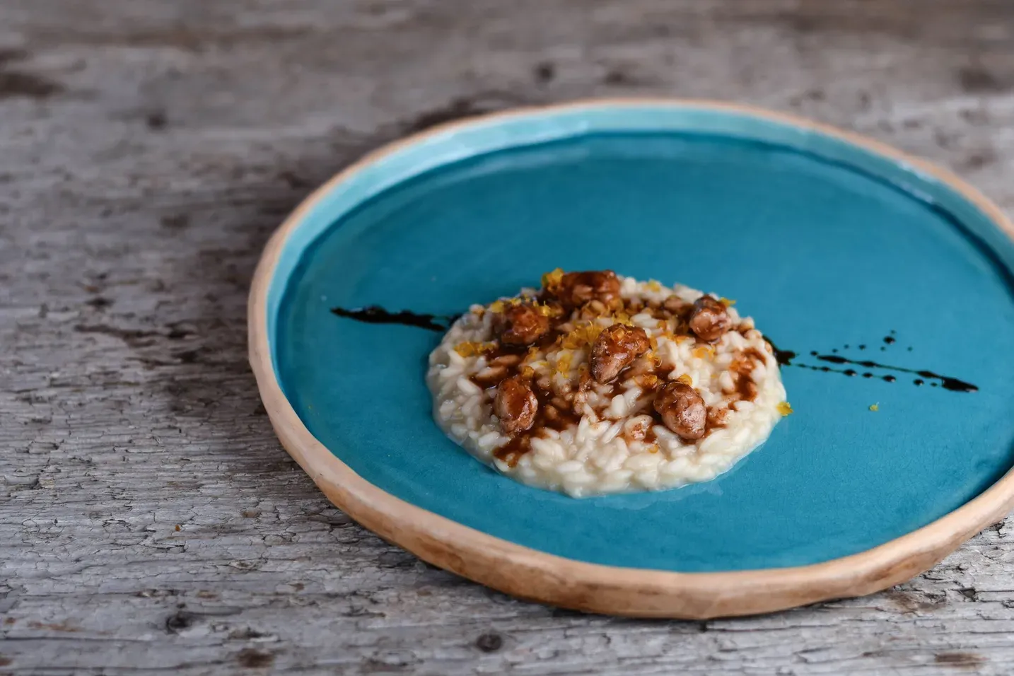 Kremiges Risotto mit gerösteten Kastanienstücken auf türkisblauem Teller, dekoriert mit dunkler Balsamico-Glasur vor rauem Holzuntergrund.