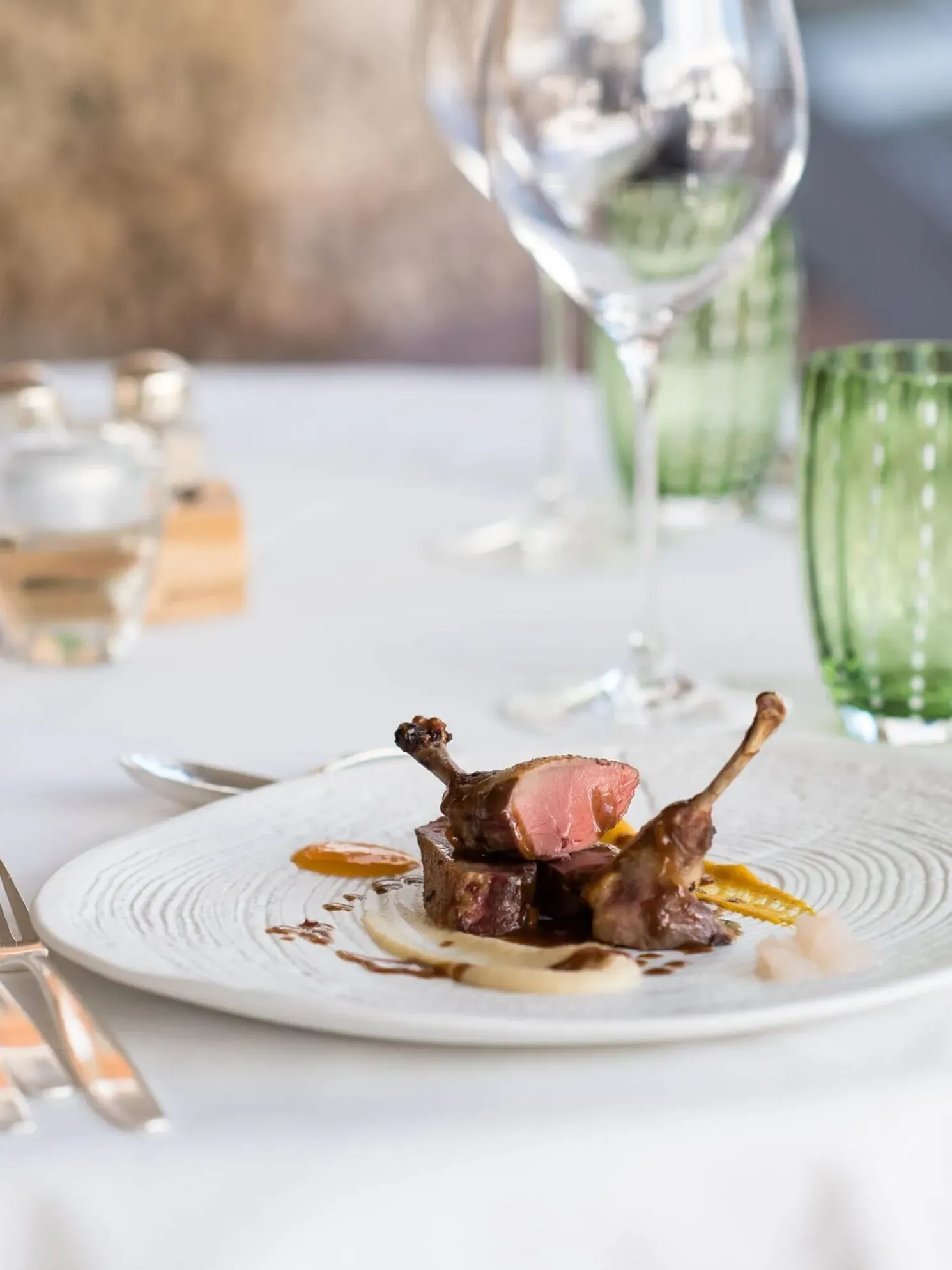 Fein angerichtetes Lammgericht mit rosa Mitte, Sauce und Beilagen auf weißem Teller; Hintergrund mit Weingläsern und grünen Gläsern auf dem Tisch.
