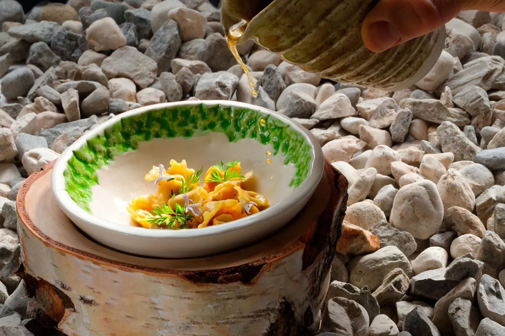 Pasta in einer weißen Schale mit grünem Rand auf einer Holzscheibe, über Kies gelegt; eine Hand gießt Sauce darüber, draußen in Alta Badia, Dolomiten.