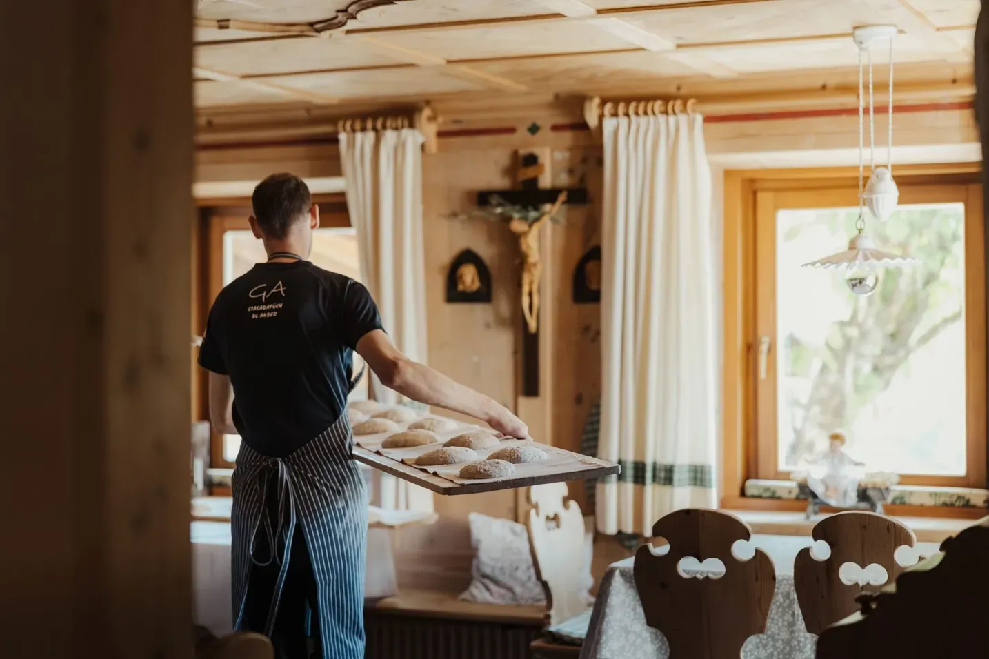 Person in schwarzem Shirt und gestreifter Schürze trägt Brötchen auf einem Backblech durch rustikales Hotelrestaurant; Holzwände, Vorhänge, Kreuz an der Wand.