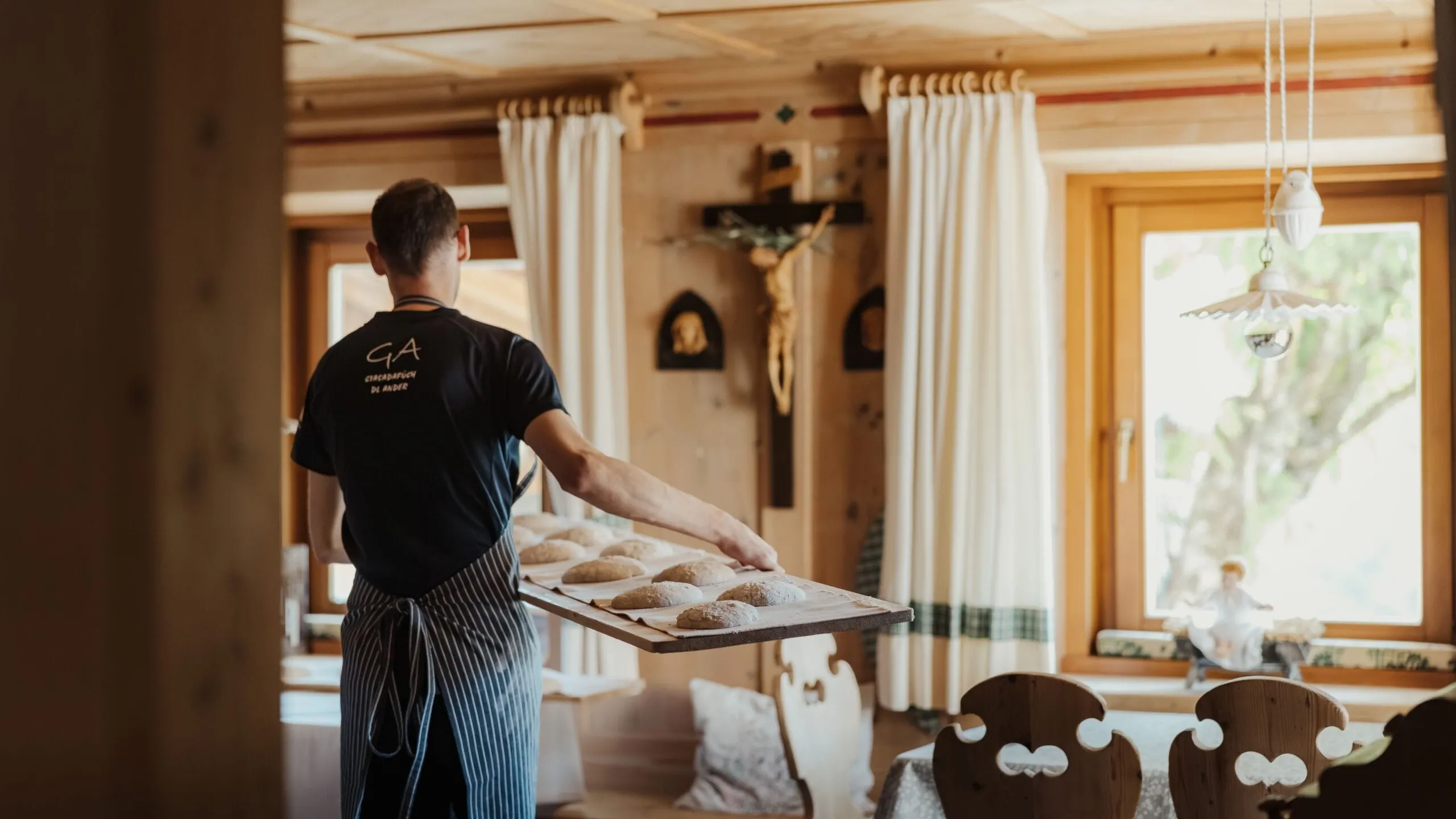 Persona di spalle in grembiule a righe tiene un vassoio con piccole pagnotte in una cucina rustica dell'Hotel Gran Ander, Badia, Dolomiti, con soffitto in legno e finestra luminosa.