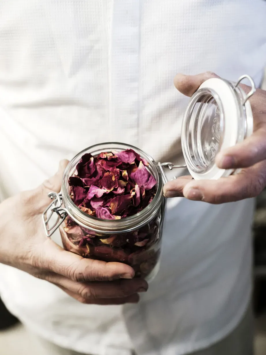 Person hält ein Glas mit getrockneten Rosenblättern; Deckel wird geöffnet, rosafarbene Blüten im Glas sichtbar.