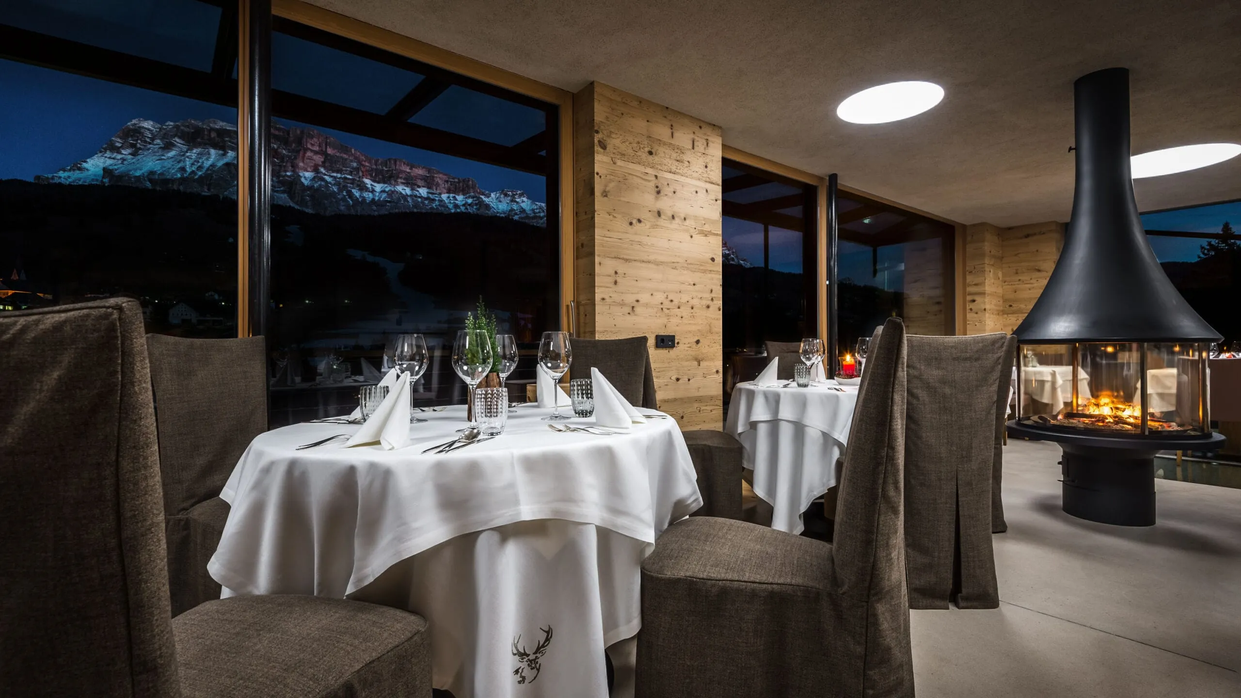 Ristorante elegante con tavoli rotondi, tovaglie bianche, tovaglioli piegati e calici; camino acceso e grandi vetrate con vista notturna sulle Dolomiti.