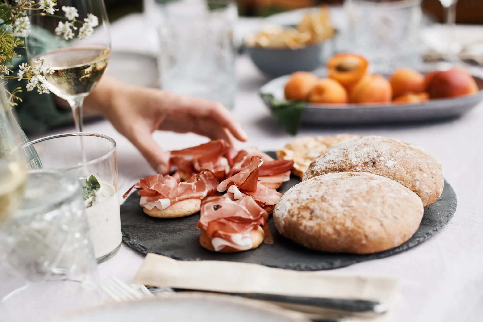 Eine Hand legt Prosciutto auf belegte Brötchen auf einer Servierplatte; daneben zwei runde Brötchen. Hintergrund: Obstkorb und Gläser Wein.