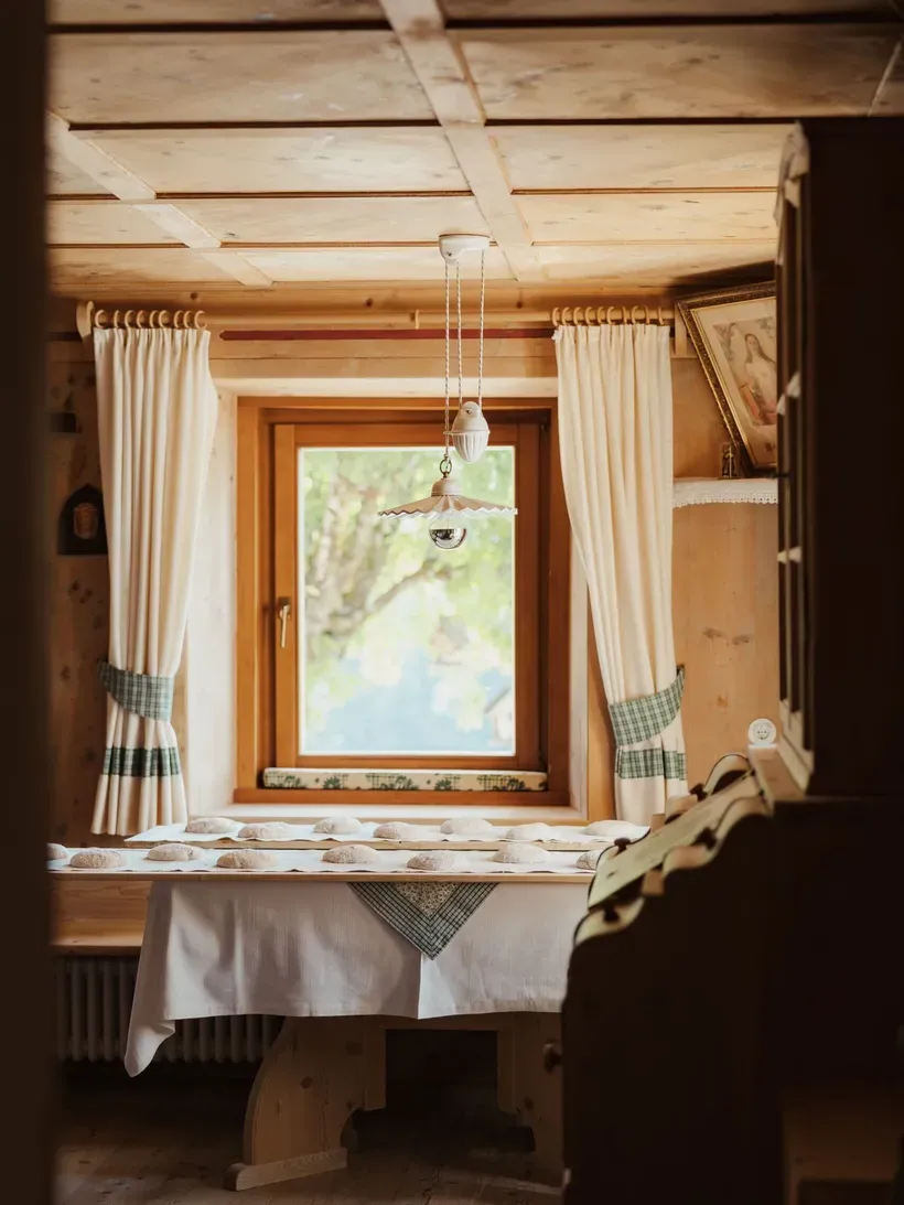 Rustikales Hotelzimmer im Hotel Gran Ander in Badia, Alta Badia: Holzdecke, Fenster mit weißen Vorhängen, Tisch mit Brotlaiben und Pendellampe.