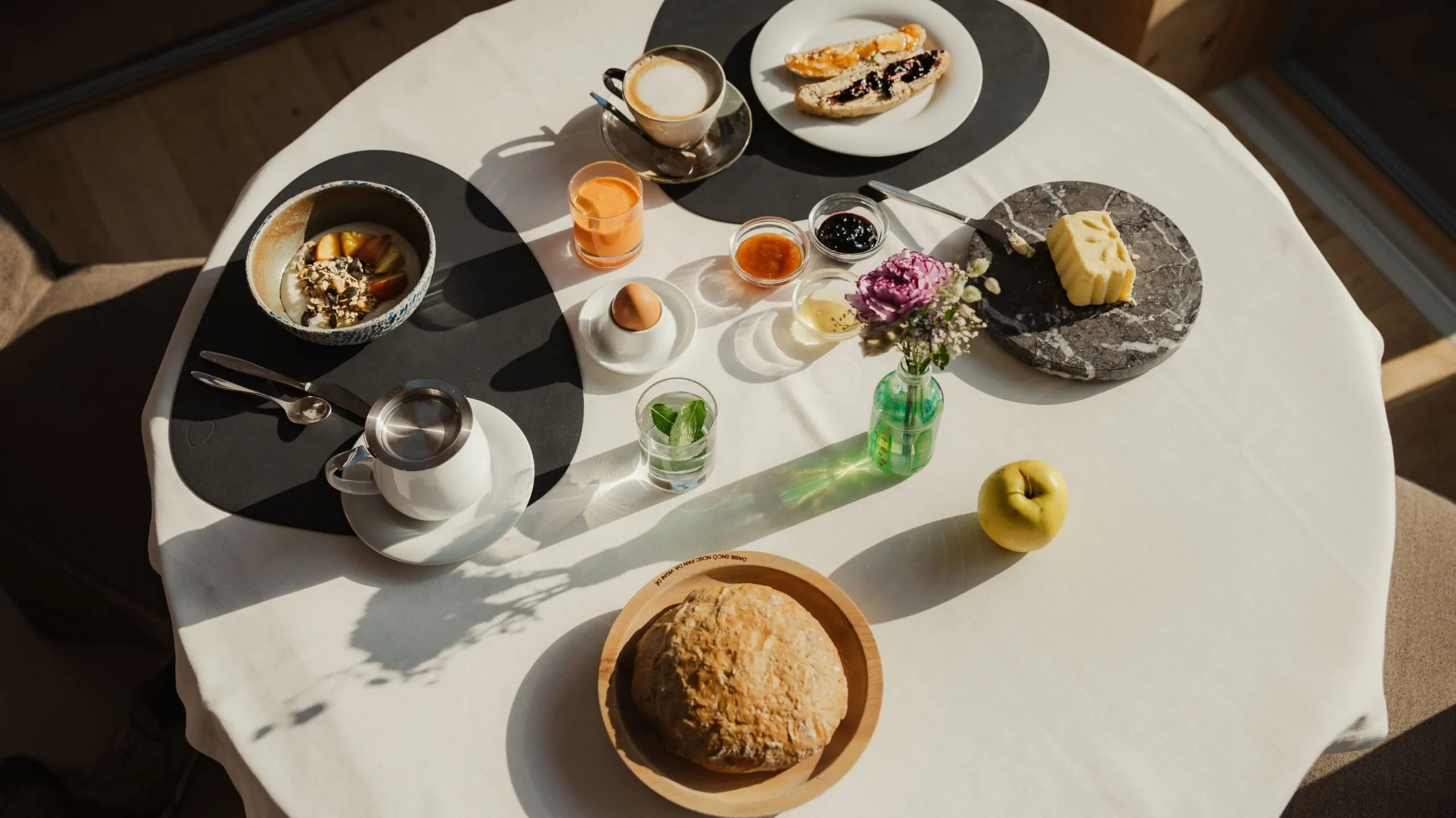 Tavolo rotondo apparecchiato per colazione: pane in ciotola, uovo, caffè, succo d'arancia, marmellata, burro, formaggio, mela e piccoli fiori.