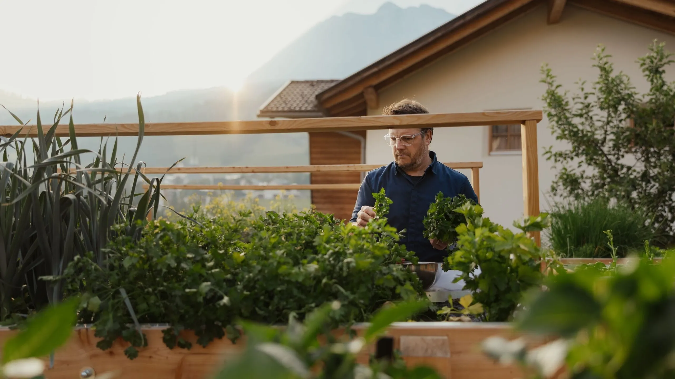 Una persona con occhiali protettivi lavora tra piante in un giardino pensile in legno, con montagne e cielo al tramonto sullo sfondo.