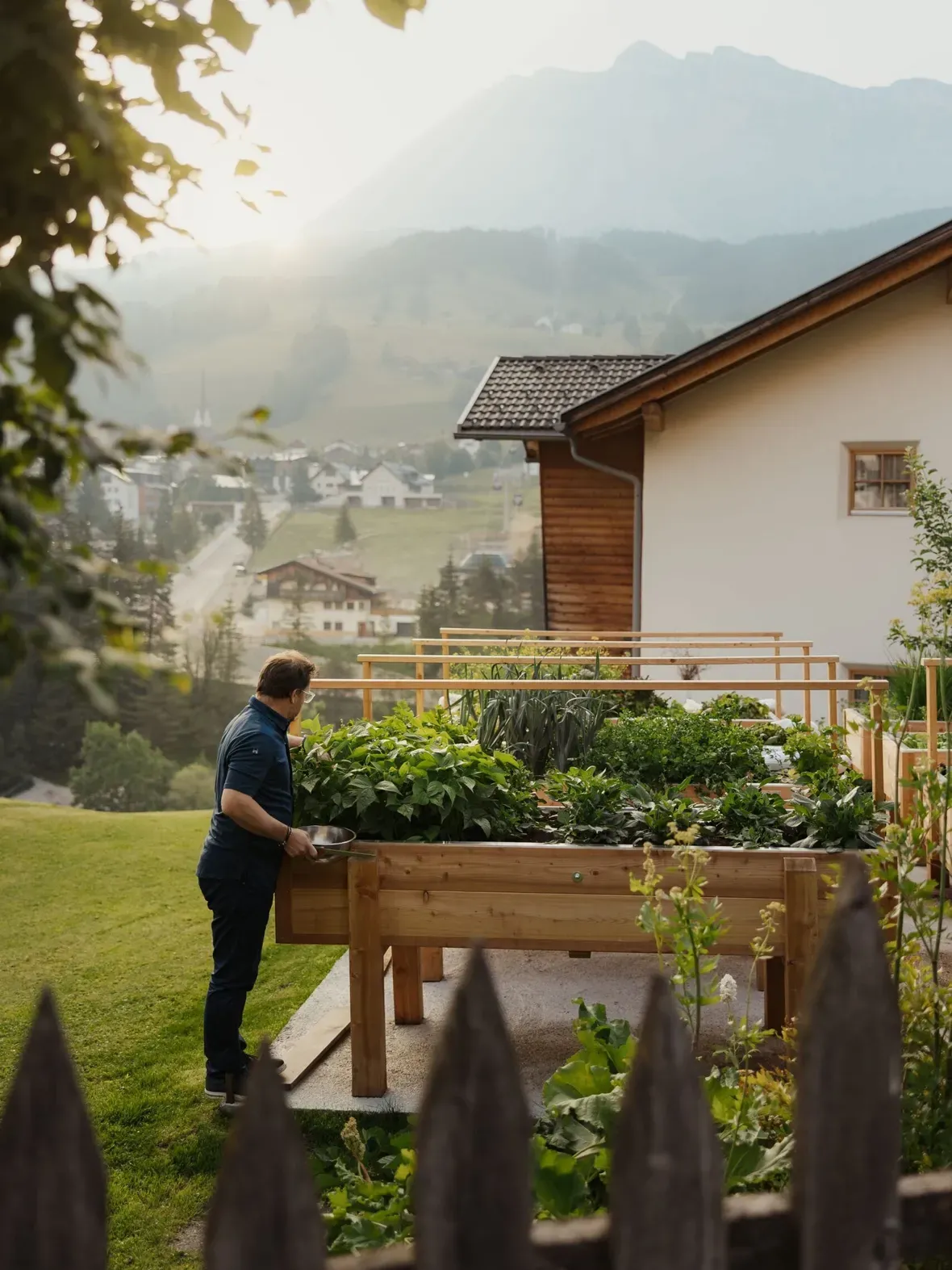 Eine Person in dunklem Shirt pflegt ein hölzernes Hochbeet im Garten eines Hauses in Alta Badia, Dolomiten; im Hintergrund Berge und ein Dorf.