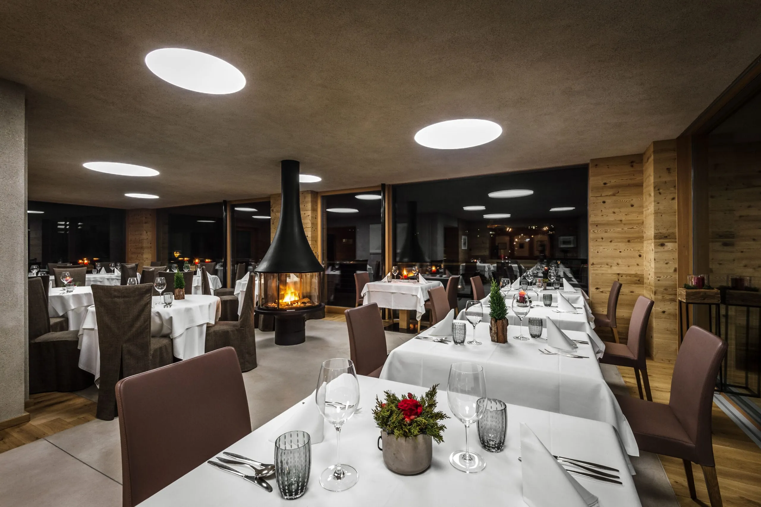 Hotel Gran Ander, Badia: heller Speisesaal mit weiß gedeckten Tischen, zentralem Kamin, Holzwandverkleidung und runden Deckenleuchten.