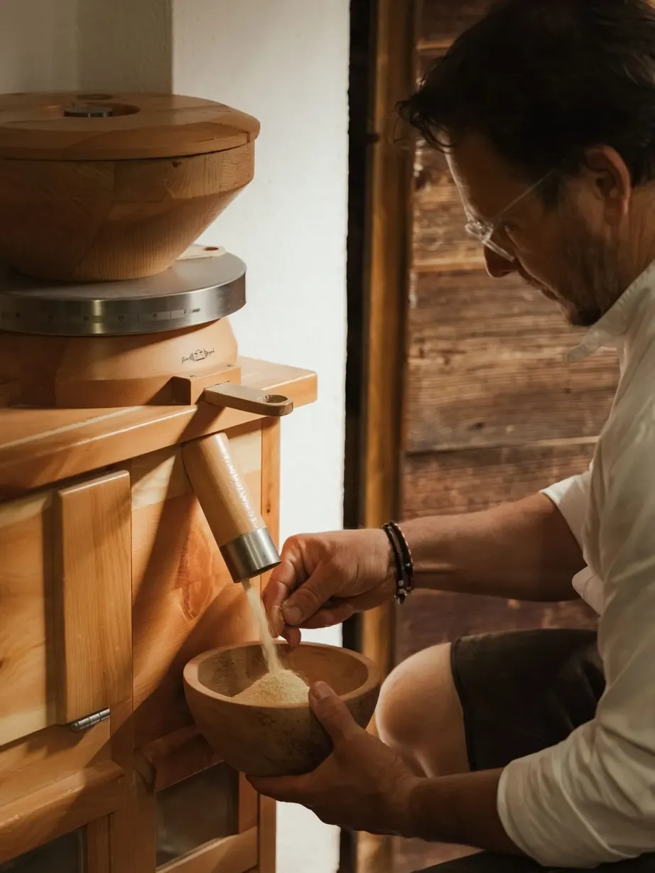 Person bedient eine traditionelle Holzmühle, mahlt Körner und fängt das Mehl in einer Holschale auf; warmes Holzambiente im Innenraum.