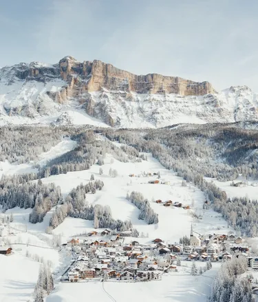 Paesaggio in Alta Badia, Dolomiti: villaggio innevato ai piedi di maestose cime rocciose, boschi innevati e chalet alpini sotto un cielo azzurro.