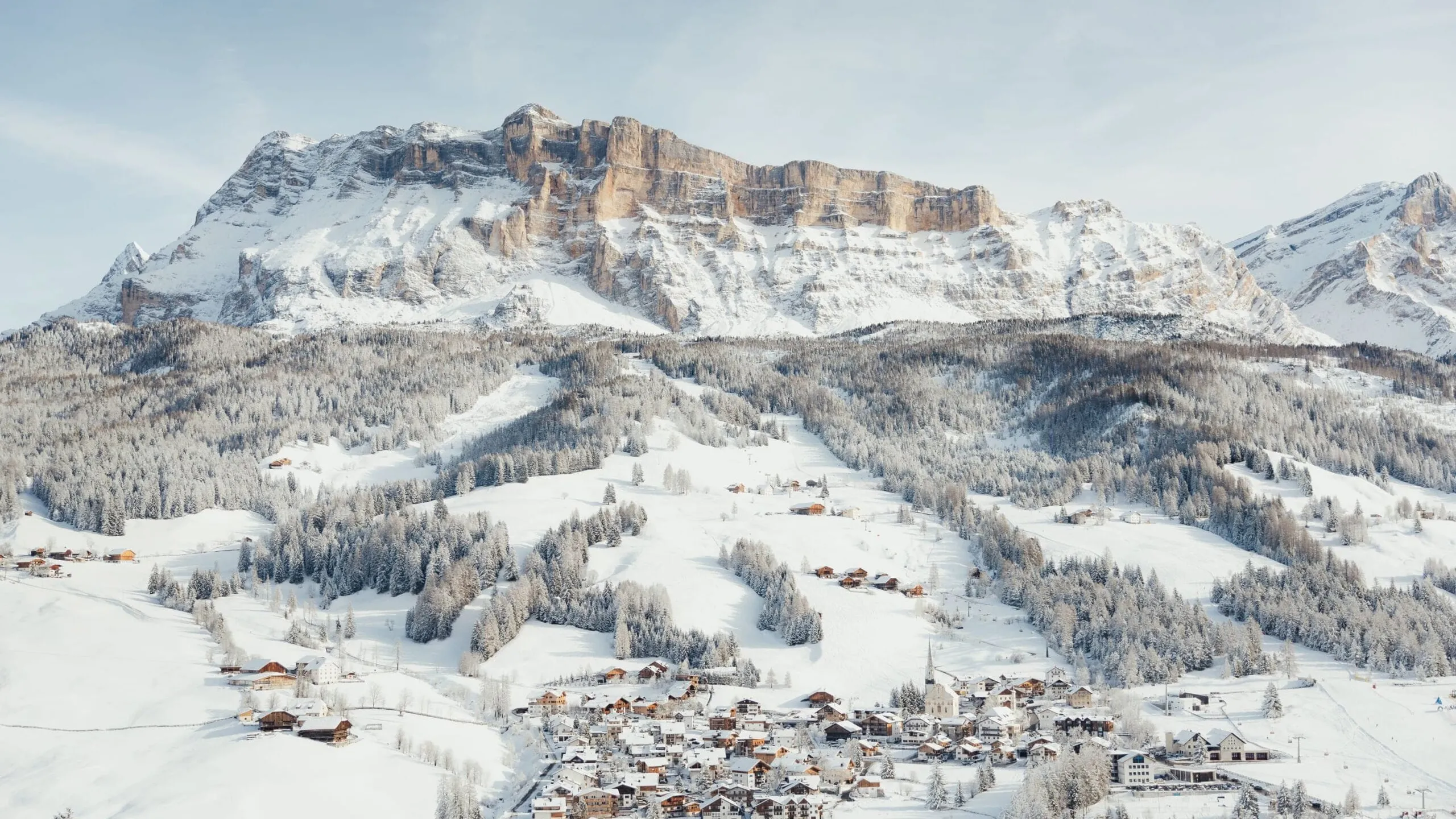Winterlandschaft in Alta Badia, Dolomiten: verschneites Dorf am Fuße einer Bergkette, von Schnee bedeckte Hänge und Nadelwälder.