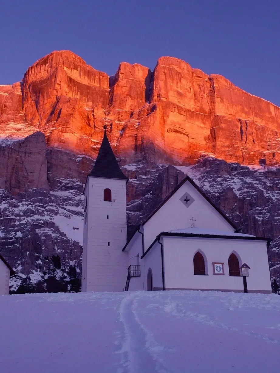 In Alta Badia, Dolomiten: Weiße Kirche mit Turm und Nebengebäude vor schneebedecktem Hang, rote Abendsonne färbt die Felswände im Hintergrund.