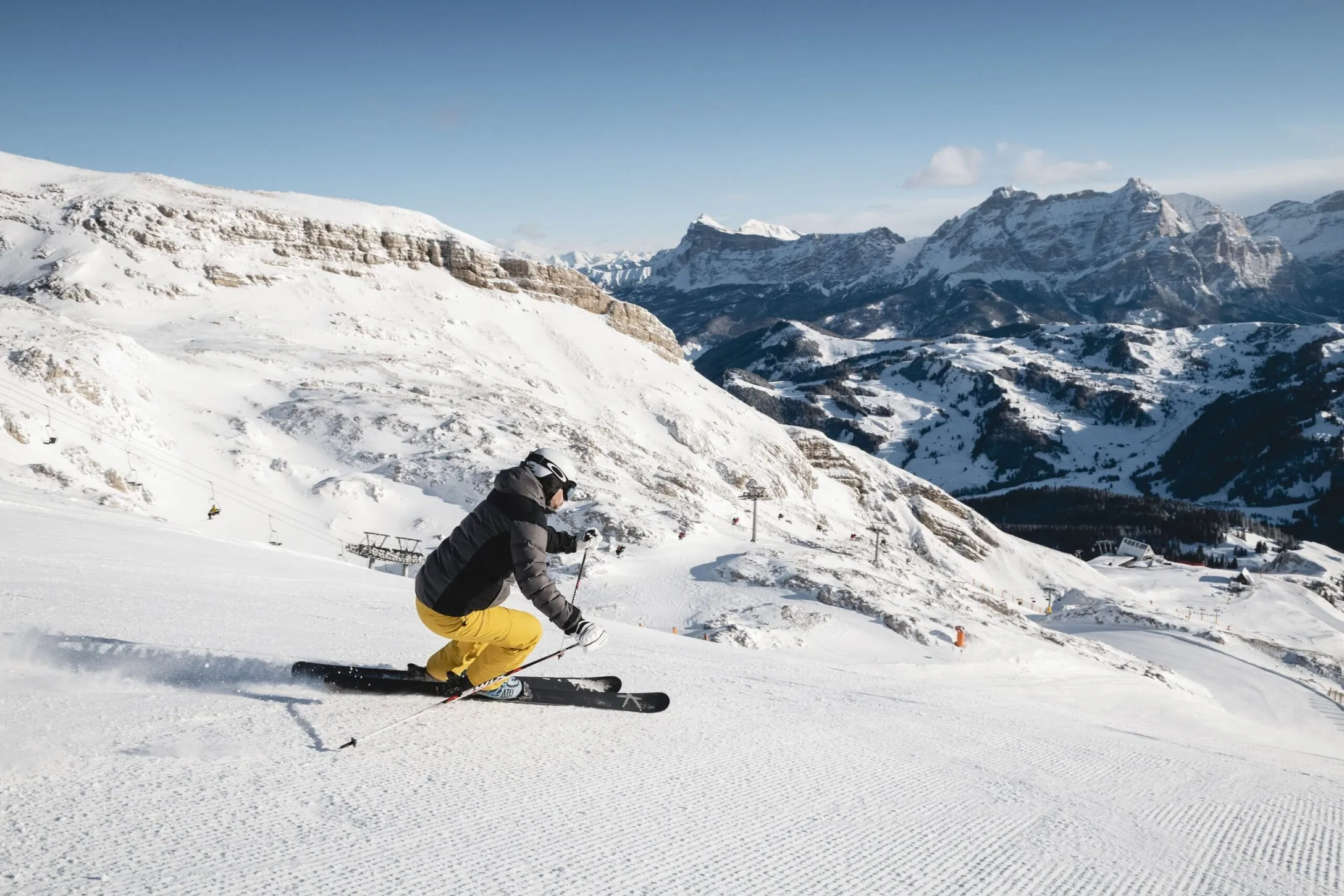 Skier wearing a gray-black jacket and bright yellow pants skis down a snowy slope in Alta Badia, Dolomites, with rugged mountains in the background.
