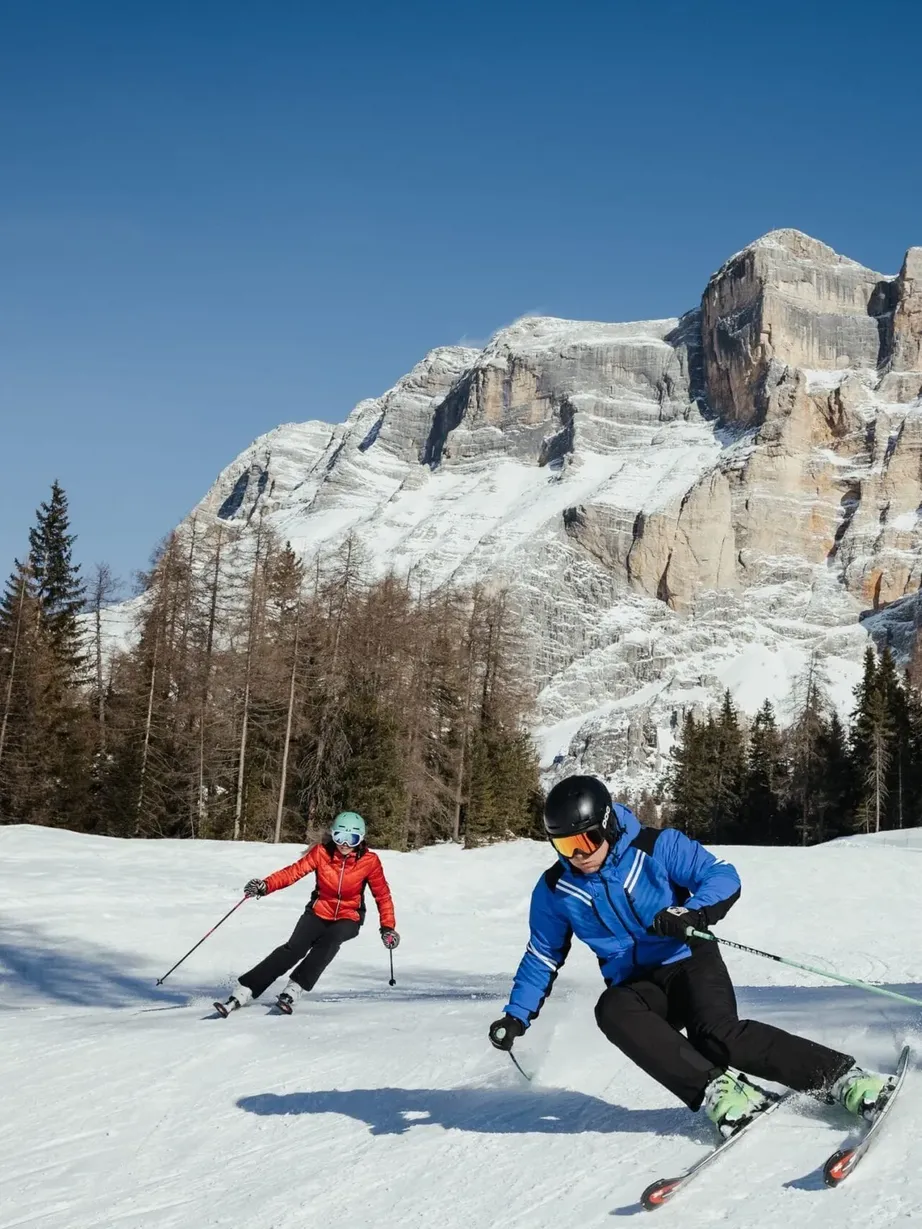 Two skiers in blue and red jackets descend a snowy slope in Alta Badia, Dolomites, with pine trees, a snow-capped mountain backdrop, and red safety fencing.