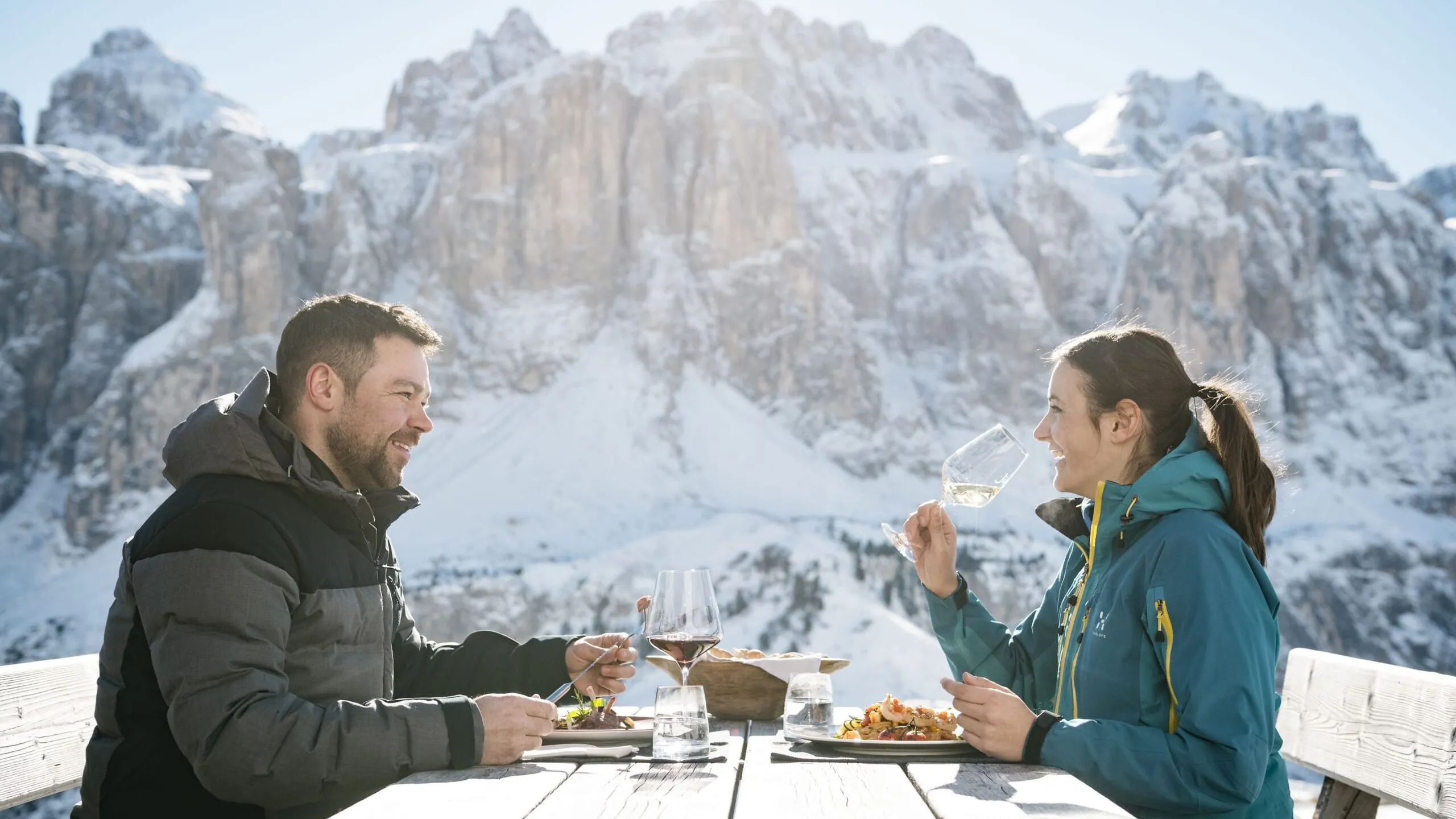 Two people dining outdoors at a wooden table in Alta Badia, Dolomites, enjoying a meal and wine with snow-capped peaks in the background.