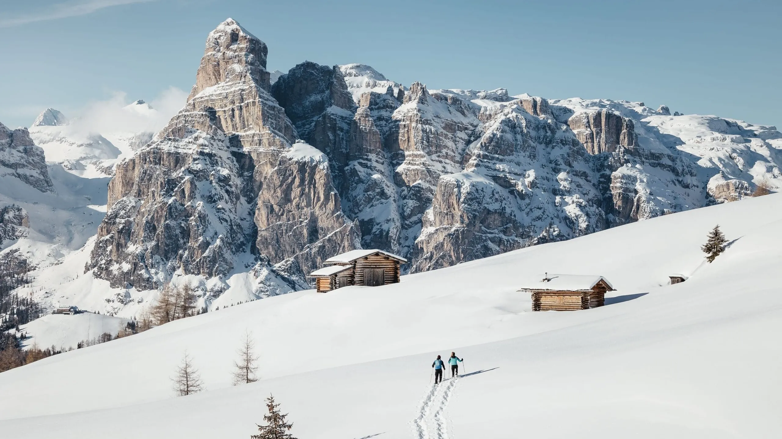 Snowy Alta Badia Dolomites landscape with jagged, snow-dusted peaks, rustic wooden huts on a gentle slope, and two hikers along a snowy trail.