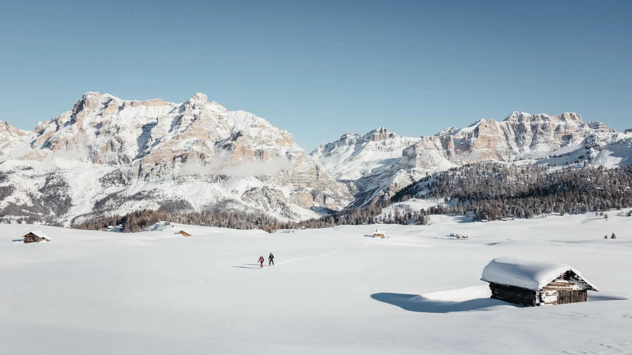 Snowy Alta Badia valley with towering Dolomites, small wooden huts, and two skiers crossing a wide, blanketed field under a clear blue sky.