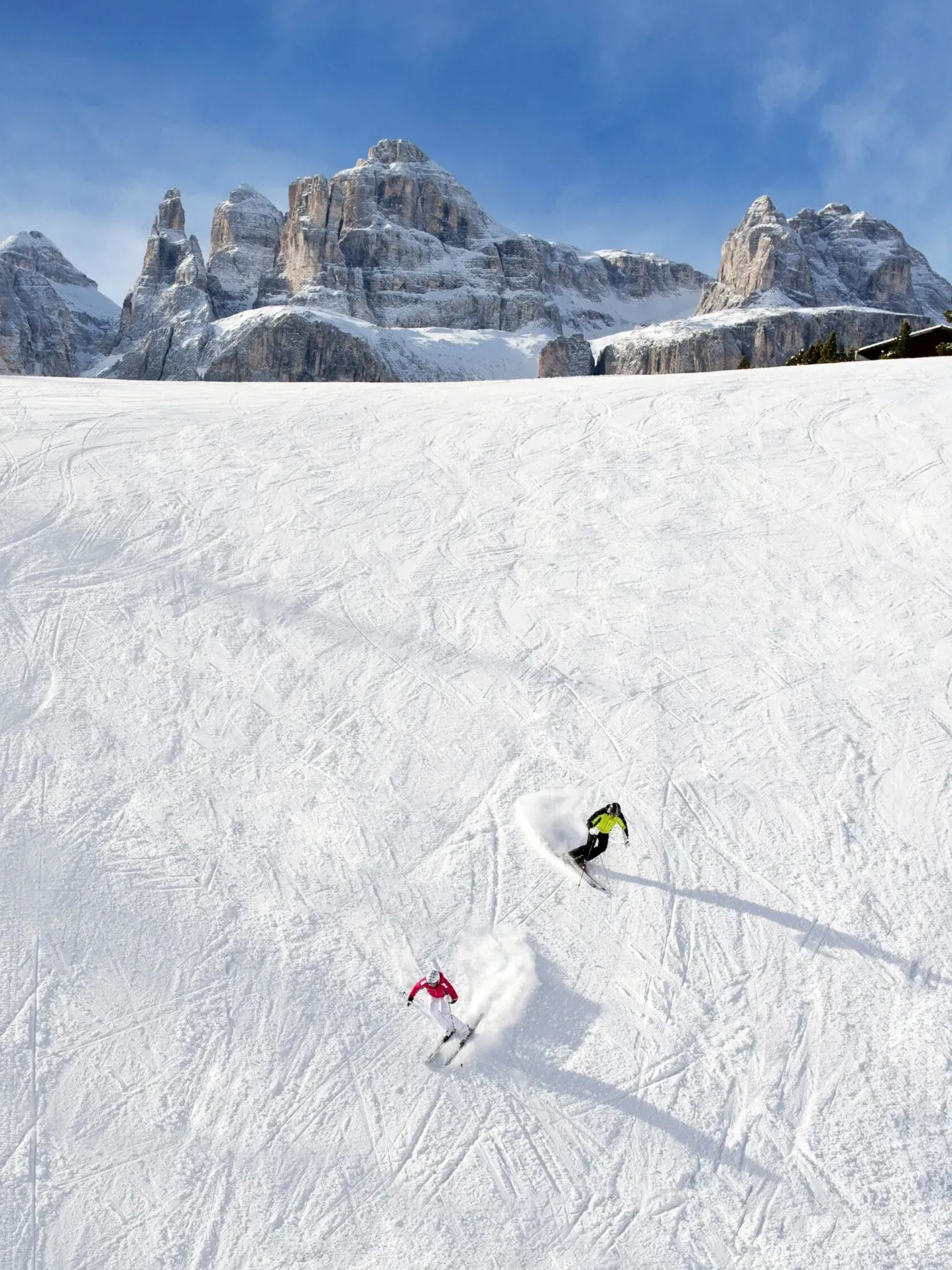 Two skiers, one in red and one in lime-green, descend a snowy slope beneath jagged Dolomite peaks and a blue sky.