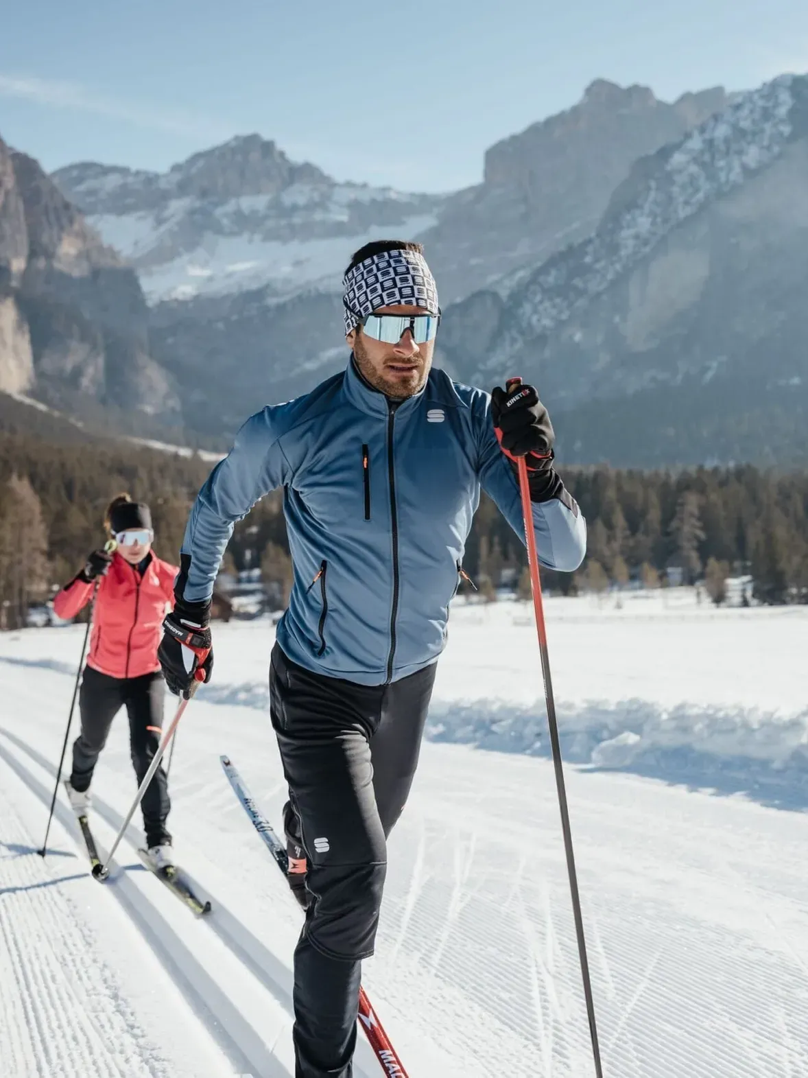 Two cross-country skiers on a groomed snow trail in Alta Badia, Dolomites, with trees and mountains; the skier in a blue jacket wears goggles.